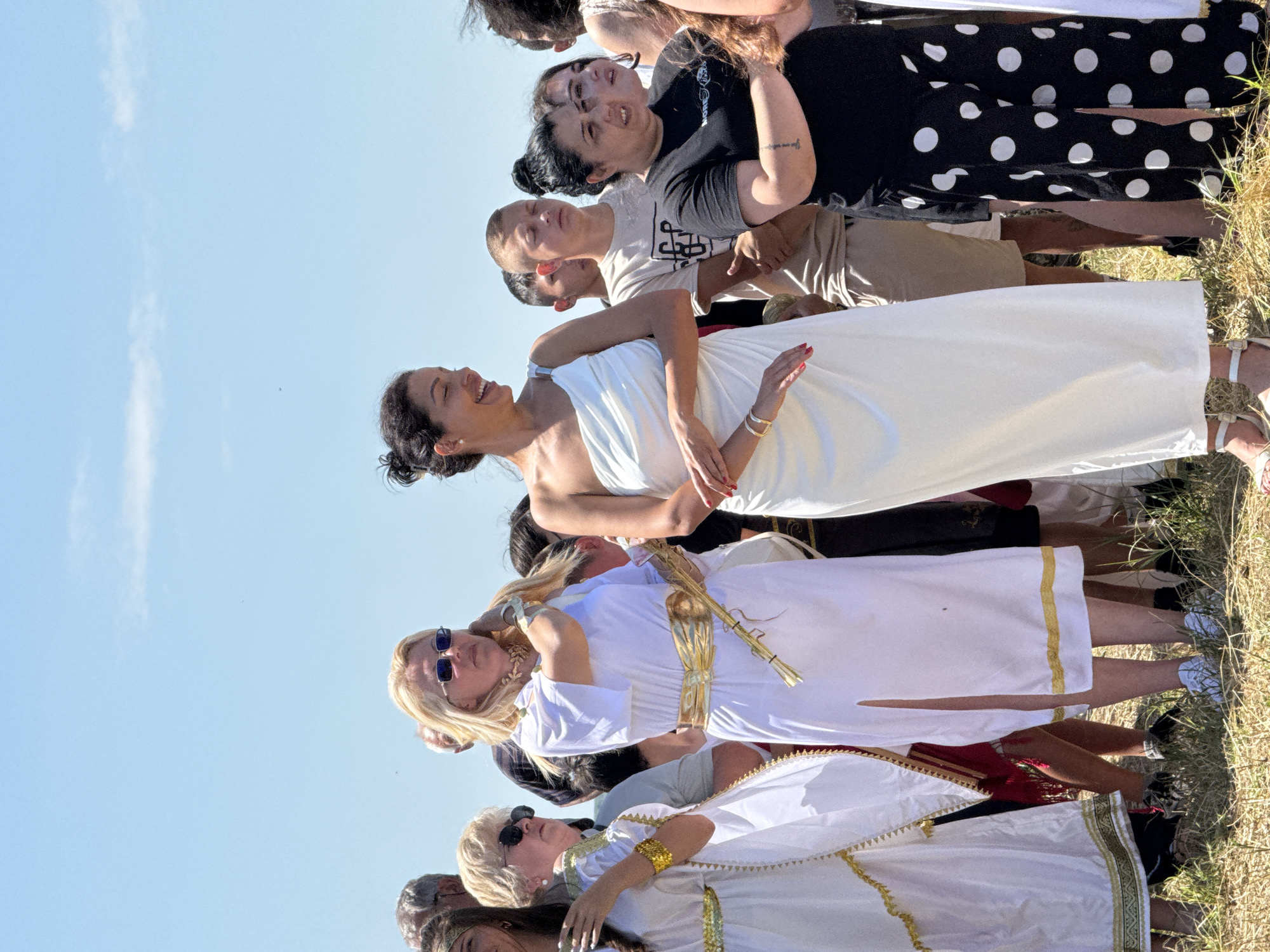This image depicts a group of individuals standing outdoors under a clear, light blue sky in Gigen, Bulgaria. The ground beneath them consists of dry, sparse yellowish-brown grass and dirt. The bright sunlight suggests midday.
In the foreground, a diverse group of approximately twelve visible individuals, primarily adults, are assembled. Several participants are dressed in white garments with gold accents, indicative of classical antiquity attire.
Specifically, from left to right:
1. A woman with dark hair and a gold-trimmed white dress is partially visible on the far left, wearing a gold armband.
2. Adjacent to her, an older woman with blonde hair, wearing dark sunglasses, a grey top, and white pants, is partially visible.
3. The central left figure is a blonde woman, with hair styled to her shoulders, wearing dark sunglasses and a white, knee-length tunic dress with a prominent gold belt. She has a gold choker necklace and is seen with her right hand raised towards her temple or hair. Her feet are partially visible, wearing white sneakers.
4. To her right, an older man with short grey hair is visible behind her.
5. The central figure is a woman with dark hair pulled into a bun, smiling broadly and looking downwards and to her left. She wears a white, strapless, toga-like dress that extends to her ankles. A gold bracelet is visible on her left wrist, and red nail polish is present on her fingers. She wears white sandals.
6. Behind the smiling woman, two young men with short hair are visible. The one closer to the center has his eyes closed or squinting, wearing a light brown t-shirt with a graphic design.
7. To the right of the smiling woman, a woman with dark hair in a bun wears a black t-shirt and black pants with white polka dots. A tattoo is visible on her right forearm. She has her arms crossed or partially obscured.
8. Further to the right, another woman with dark hair is visible, looking to her right.
9. On the far right, a person with curly dark hair and another individual in a white garment are partially visible.
The individuals are standing upright, mostly facing forward or slightly to their left, suggesting they are observing an event or presentation outside the frame of the image.