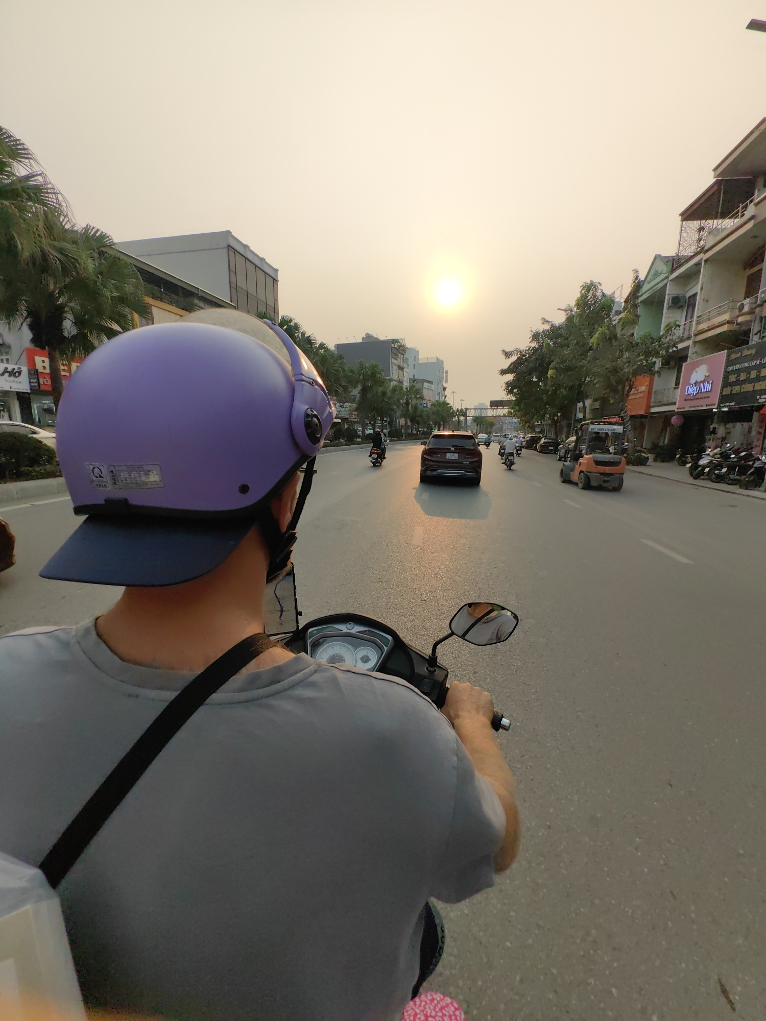 A street-level, eye-level, outdoor shot depicts a person riding a scooter through Ha Lam Ward, Vietnam. The scooter rider, wearing a lavender helmet with a dark blue brim and a gray t-shirt, is facing away from the camera, showing the back of their head and shoulders. A black strap crosses their torso. The view is over the rider's shoulder, showing the street ahead.
The street is lined with buildings, some with awnings, and trees. Traffic includes cars, motorbikes, and a forklift. The sun is visible in the distance, creating a hazy, golden light. Street signs and Vietnamese script are visible on the buildings.
