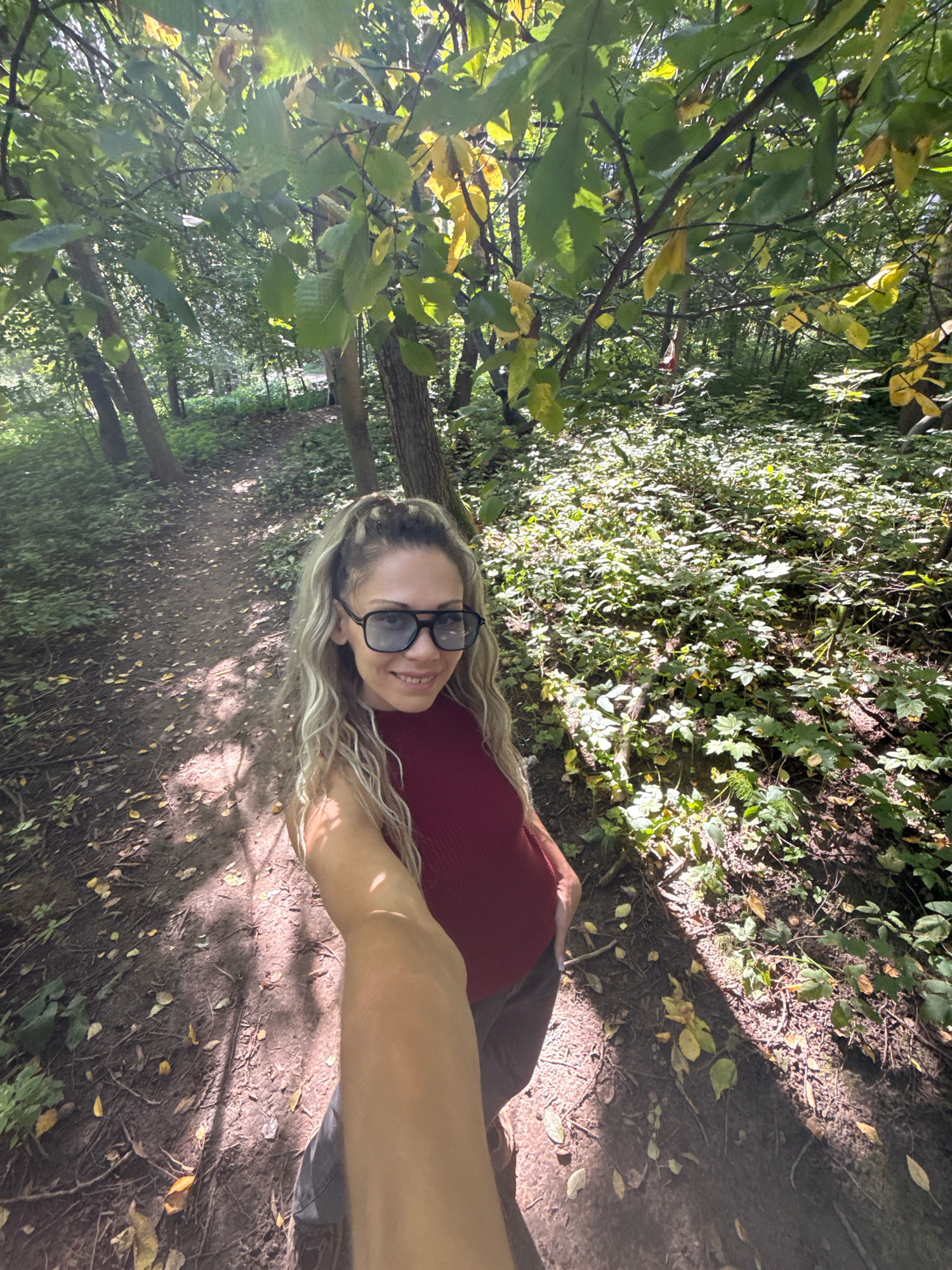 A woman is taking a selfie in a forest. She has long blonde hair, is wearing large sunglasses, and a red top. She is standing on a dirt path that is covered with fallen leaves. The sun is shining through the trees, creating dappled light on the ground. The forest is dense with green foliage. This scene is located in the city of Rusanovo, Russia.