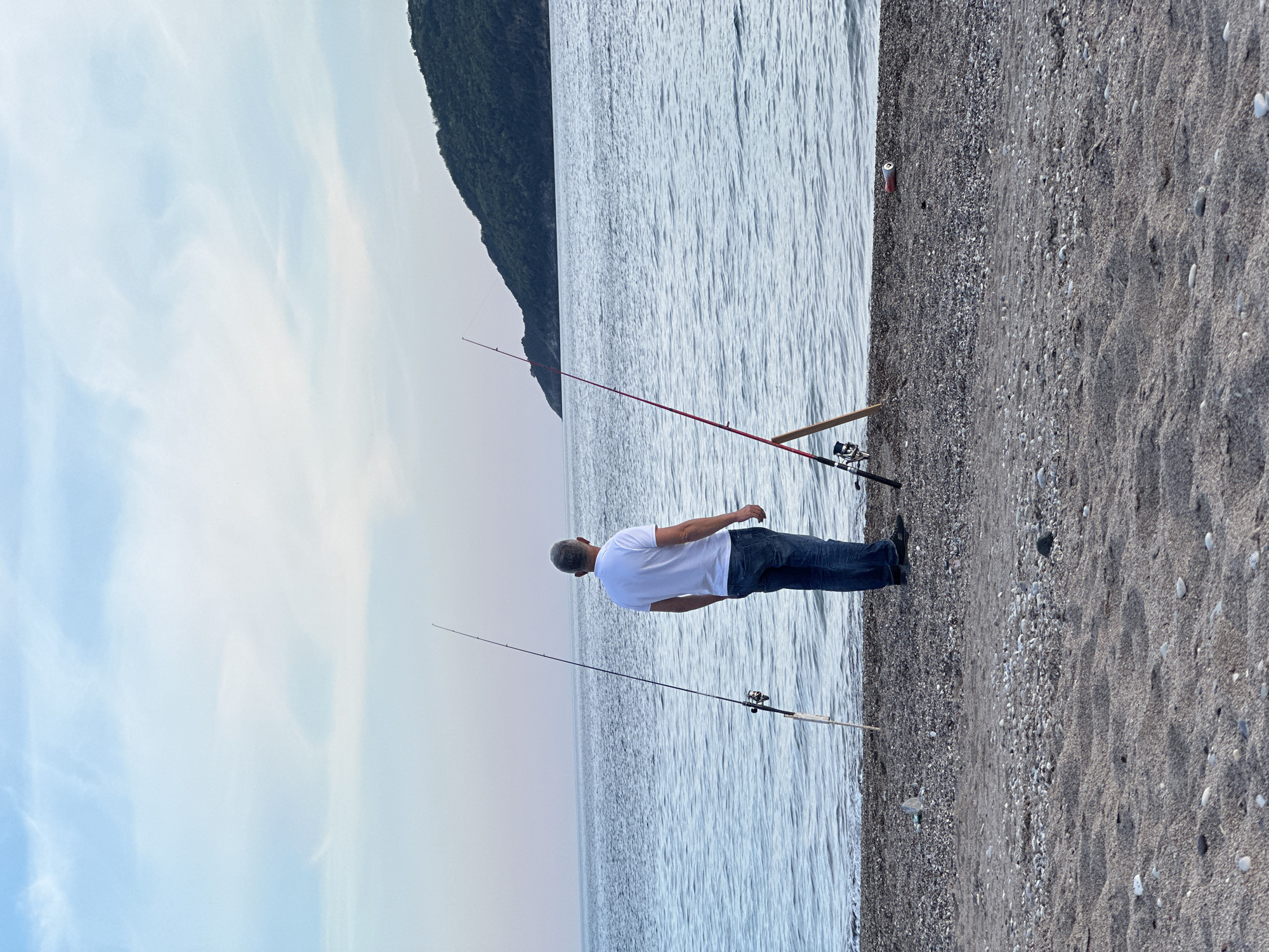 A man stands on a sand and gravel beach in Budva, Montenegro, fishing with three fishing rods. He is wearing a white shirt and dark blue jeans. The sky is cloudy. There is a mountain in the background. A red soda can sits on the beach behind the rods.