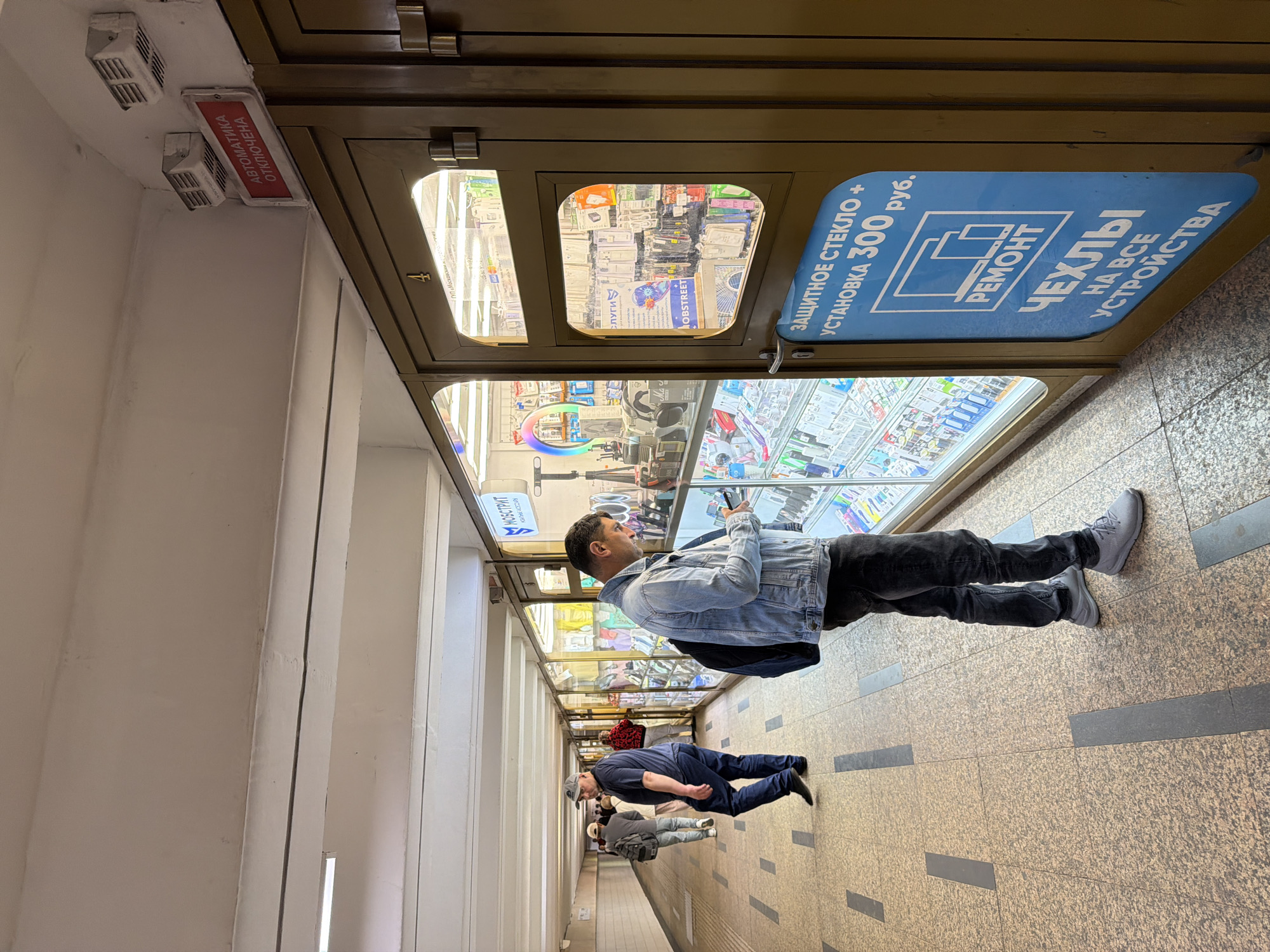 Here is a detailed description of the image:
The image shows an interior view of a possibly underground pedestrian walkway or subway station. The walls and ceiling are white with structured patterns. There is a line of shops or vendors along the right side. The walkway has tiled flooring with dark gray rectangular insets.
In the foreground, a man in a denim jacket and dark pants is standing looking at the displays inside one of the shops. Further down the walkway, there are other people walking in the same direction.
Signs and advertisements are displayed on the vendor stalls, one blue sign advertises protective glass and screen repair services. Text and other signage are in Cyrillic script, indicating that the location is likely in Russia or another country where Cyrillic is used.
