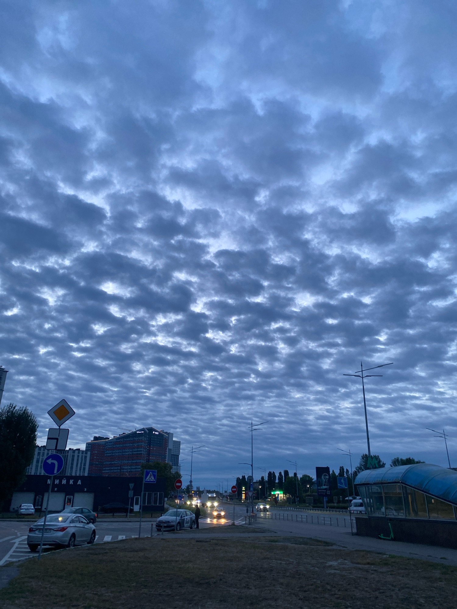The image depicts a street scene in an urban environment under a cloudy sky. The sky is filled with numerous small, puffy clouds that are arranged in a somewhat uniform pattern. The lighting suggests it is either early morning or late evening, as the light is dim.
In the foreground, there is a paved road with a grassy verge. Several cars are visible on the road, some moving and others parked. A silver sedan is in the left lane, and a dark-colored car is in the right lane, with a person standing next to it. A green electric scooter is leaning against the side of a structure on the right.
Further down the road, there are streetlights and traffic lights. Buildings of varying heights are visible in the background, including a multi-story building with a dark facade and windows, and another building with red and blue architectural elements. A sign with Cyrillic lettering, "МИЙКА", indicating a car wash, is present on one of the buildings. A blue subway entrance is visible on the right side of the image. Traffic is present on the road, with headlights of approaching vehicles visible.
The overall impression is a quiet moment in a city, possibly during a transitional time of day, with a distinctive cloud formation overhead.