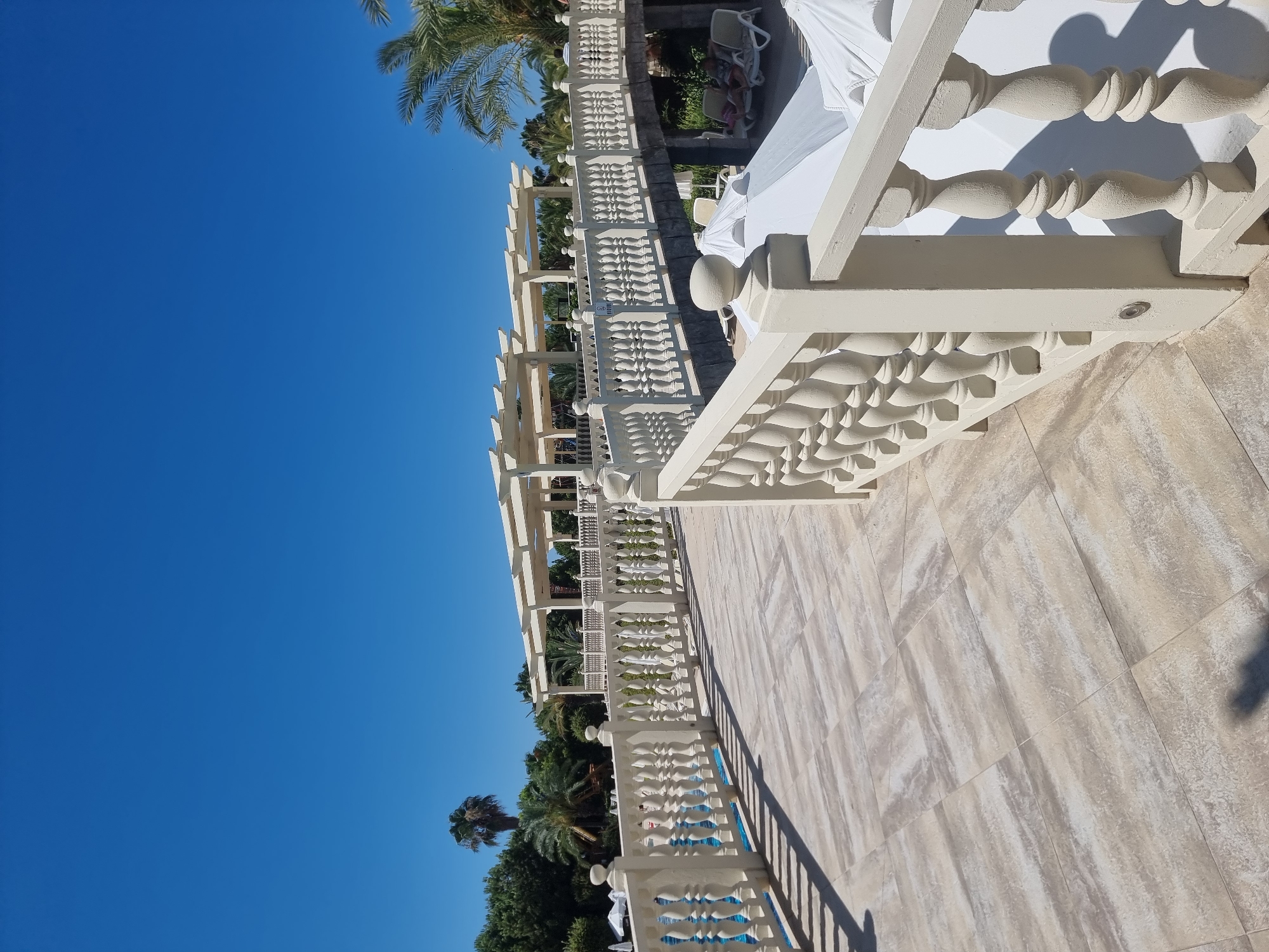 The image shows a picturesque resort setting with a clear blue sky and lush green trees. A cream-colored balustrade lines a pathway, leading towards a bridge that spans over a lower area. The pathway is paved with light-colored tiles, and shadows from the balustrade create a patterned effect. Beyond the balustrade on the left, a swimming pool with blue water is visible. The bridge has a similar cream-colored balustrade and is covered by a white pergola structure. Underneath the bridge, lounge chairs and white umbrellas suggest an area for relaxation. Palm trees are scattered throughout the landscape, adding to the tropical ambiance. The scene appears to be located in Manavgat, Turkey, a city known for its resorts and natural beauty.