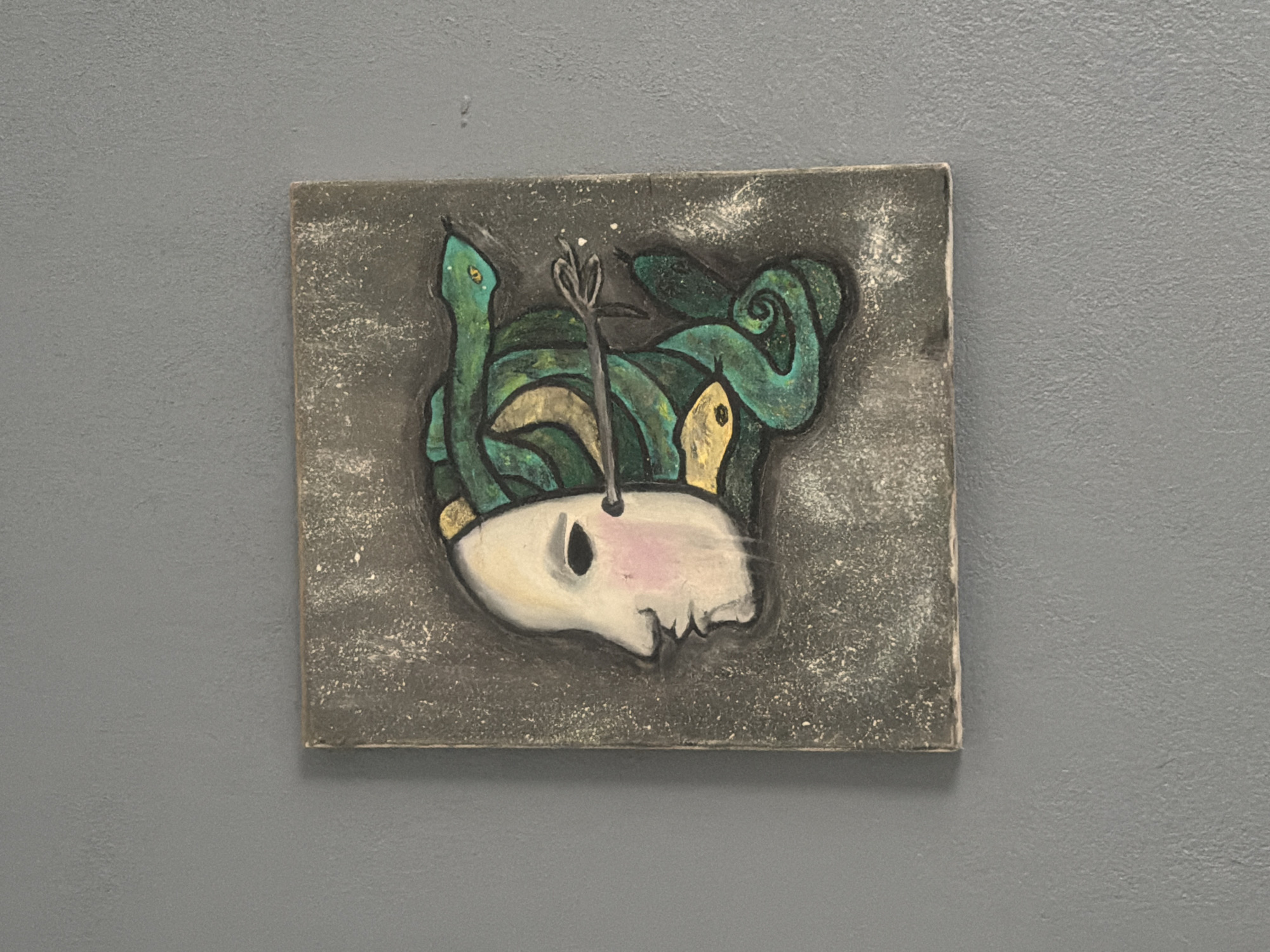 The image shows a square painting mounted on a gray wall. The painting depicts a white skull with green snakes emerging from its head, one of which is pierced by a dark, leafless branch. The background of the painting is a mottled gray color. The location is not discernable from the image.