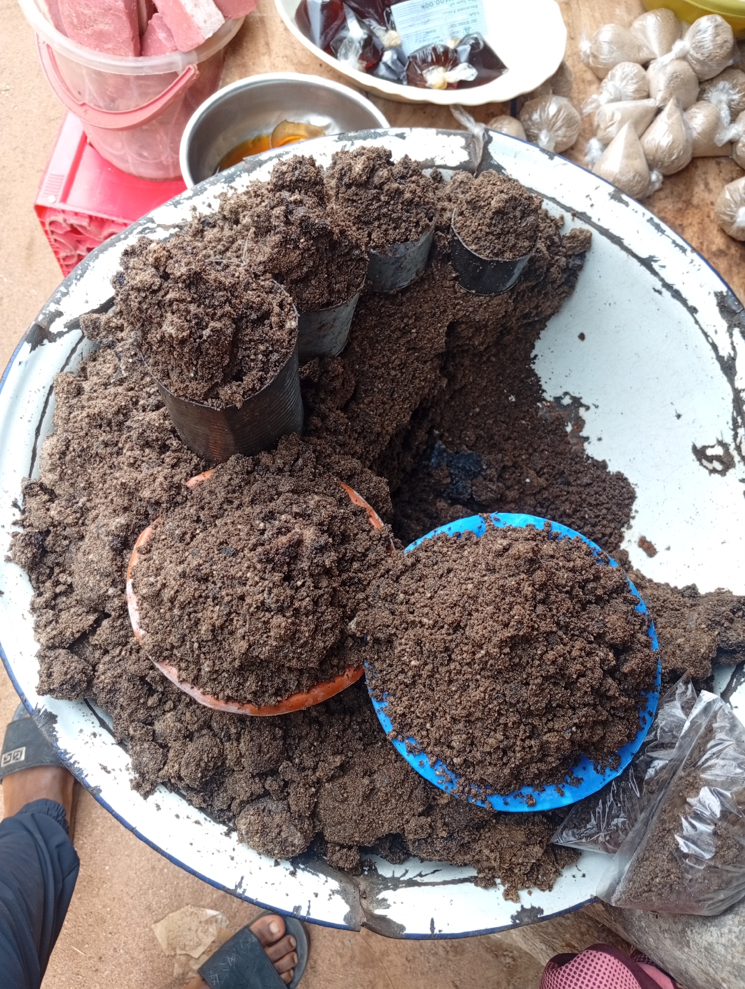 In Andaha, Nigeria, a market scene unfolds with a close-up view of a large, weathered enamel basin filled with dark brown, granular material, likely soil or a soil-based compound. Within the basin, two small, open cans are partially submerged, also filled with the same material. Two shallow bowls, one orange and one blue, are placed on top of the soil, each brimming with it. To the right of the basin, a clear plastic bag holds a portion of the material. In the background, several small, individually wrapped parcels are visible, also containing a light brown granular substance. Further back, a white bowl holds dark, jelly-like substances, and a pink bucket contains rectangular pieces of what appears to be meat or solidified foodstuff. A red plastic crate is positioned beneath the bucket. The ground beneath the display is a dusty, reddish-brown. A person's feet are visible in the lower left and bottom center, wearing black sandals. Another person's foot, wearing a pink and black shoe, is partially visible in the lower right corner. The scene suggests a retail setting for agricultural or culinary products.