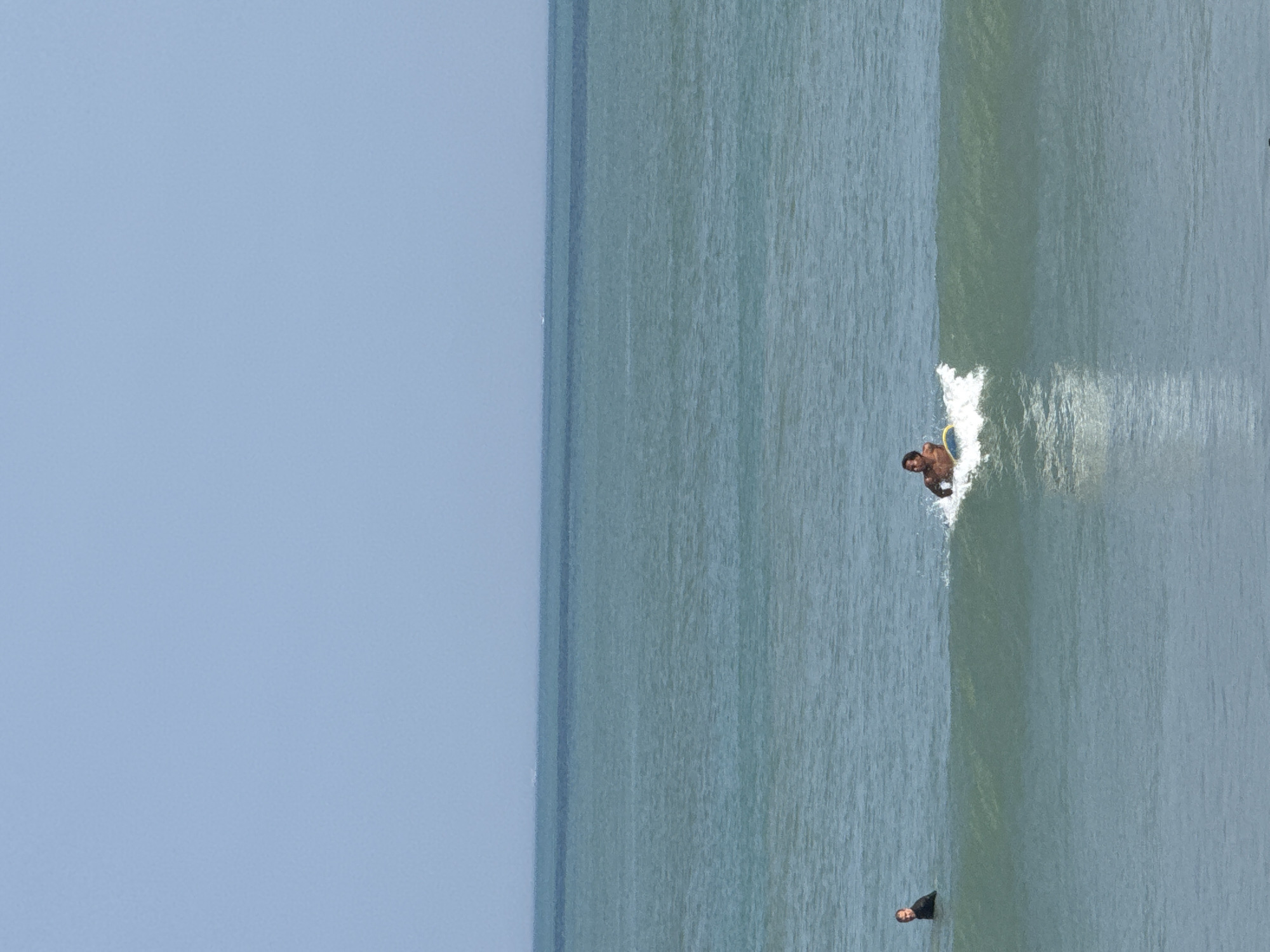 Two men are surfing in the ocean. One man is on the left, wearing a black wetsuit and looking towards the left. The other man is on the right, on a surfboard, riding a wave and looking forward. The ocean is blue-green with small waves. The sky is clear and pale blue. The image appears to be taken from a beach in Kuta, Indonesia.