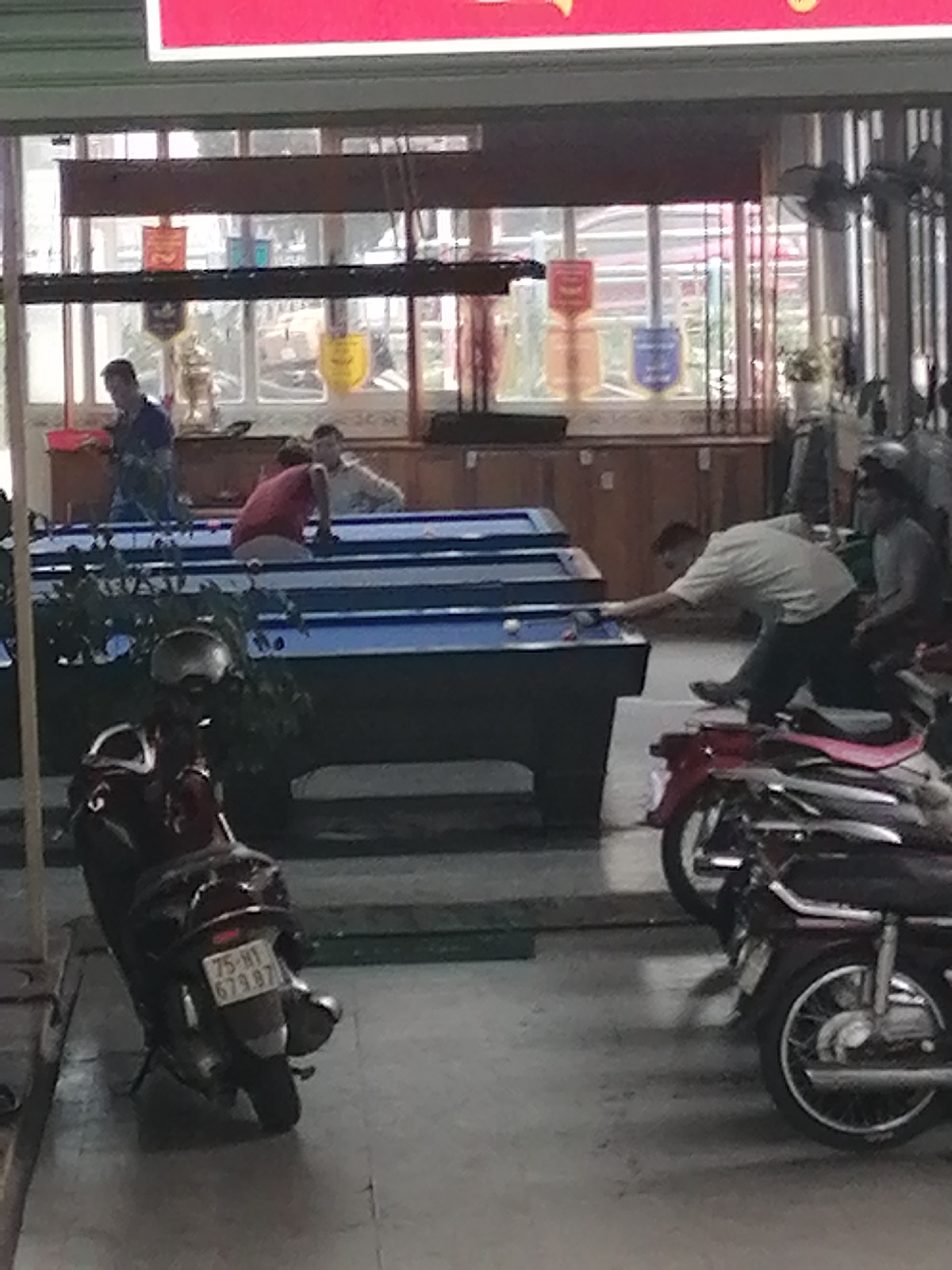 The image depicts an indoor billiard hall with several people playing pool. In the foreground, two motorcycles are parked. One motorcycle, a dark red scooter, is prominently displayed with a license plate reading "75-H1 679.87". Behind it, another motorcycle, partially obscured, is also visible.
In the midground, three billiard tables are set up. Two are covered with blue felt, and one is partially visible and appears to be the same color. People are actively engaged in playing pool. One man in a light-colored shirt is leaning over a table, appearing to take a shot. Another individual in a red shirt is seated at the edge of a table. A third person in a blue shirt is standing near a table on the left.
The background shows a row of windows with decorative banners or crests hanging on them. Some shelves with various items are also visible behind the billiard tables. The overall lighting suggests an indoor environment, possibly in a commercial establishment.
The license plate "75-H1 679.87" indicates the motorcycle is registered in Vietnam. Da Lat is a city in Vietnam known for its cooler climate and French colonial architecture, and while the image does not directly show any specific landmarks of Da Lat, the presence of a Vietnamese license plate and the general appearance of the establishment are consistent with a location in Vietnam.