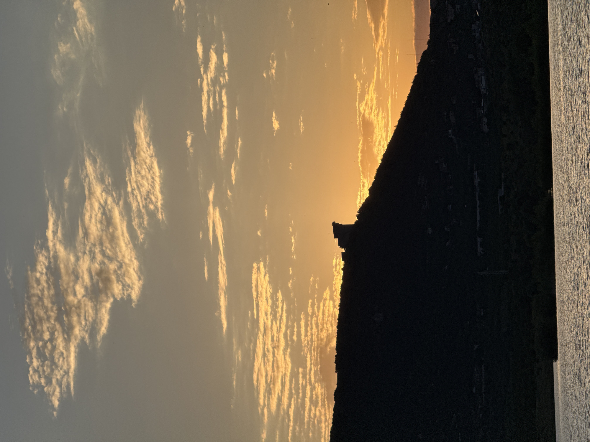 An outdoor, landscape photograph captures a silhouetted hill against a warm sunset sky. The hill, which is dark and appears to be covered in vegetation and some scattered structures, dominates the lower half of the frame. A body of water, reflecting the colors of the sunset, occupies the bottom of the image. Above the hill, the sky transitions from a pale yellow near the horizon to a soft blue at the top, with streaks of illuminated clouds scattered throughout. The overall impression is one of a serene natural scene at dusk, with the hint of civilization on the hillside. The image is taken from a low vantage point, looking across the water towards the hill. The location is not specified in the image.