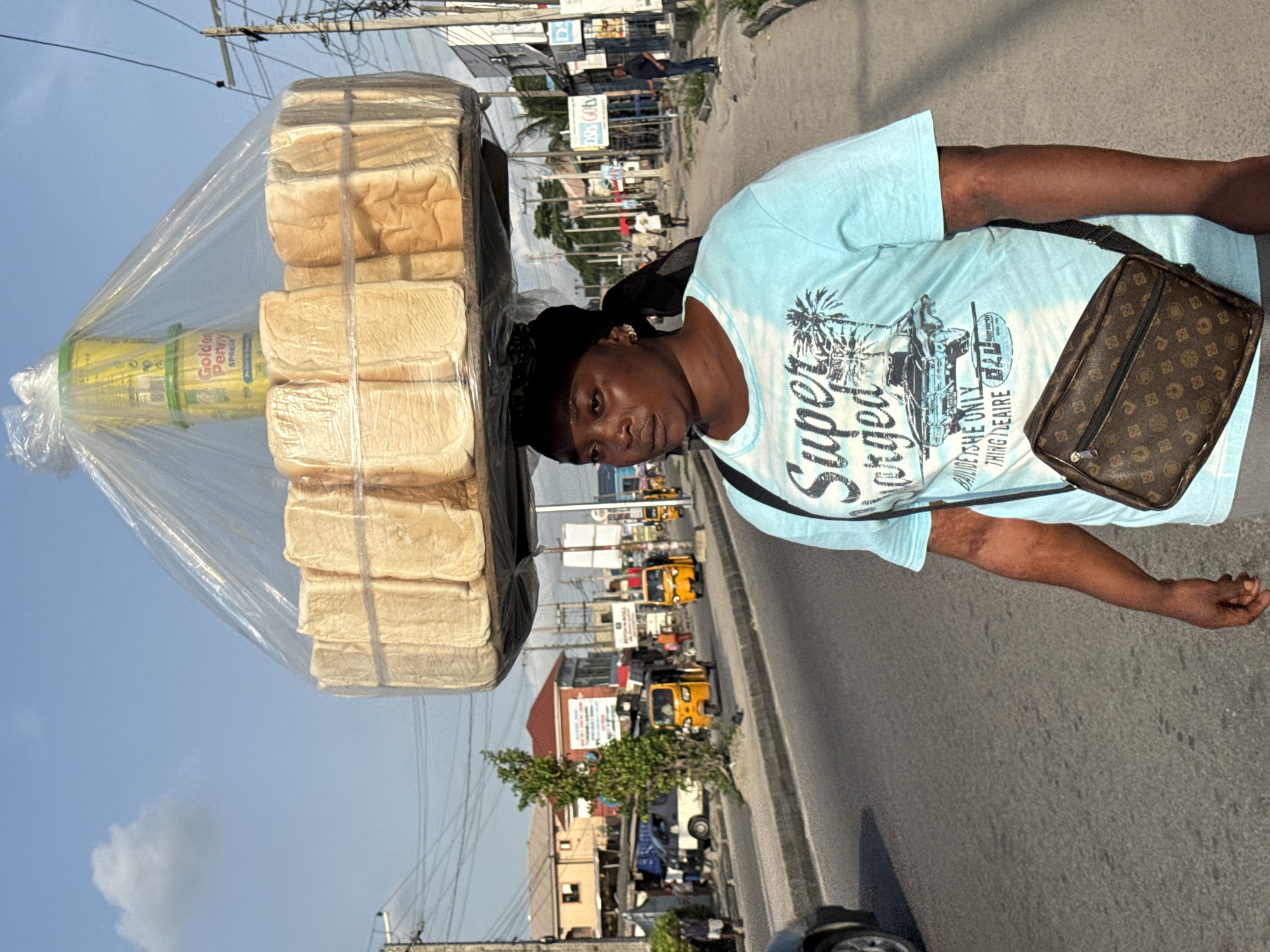 A street scene in Ado, Nigeria, shows a person standing on the side of a road. They are carrying a large stack of bread loaves in a clear plastic bag balanced on their head, along with what appears to be a container of butter. The person is wearing a light blue T-shirt, a black head covering, and a brown patterned bag across their body. The background reveals the city street with buildings, yellow three-wheeled vehicles, and other cars. Overhead power lines crisscross the sky.