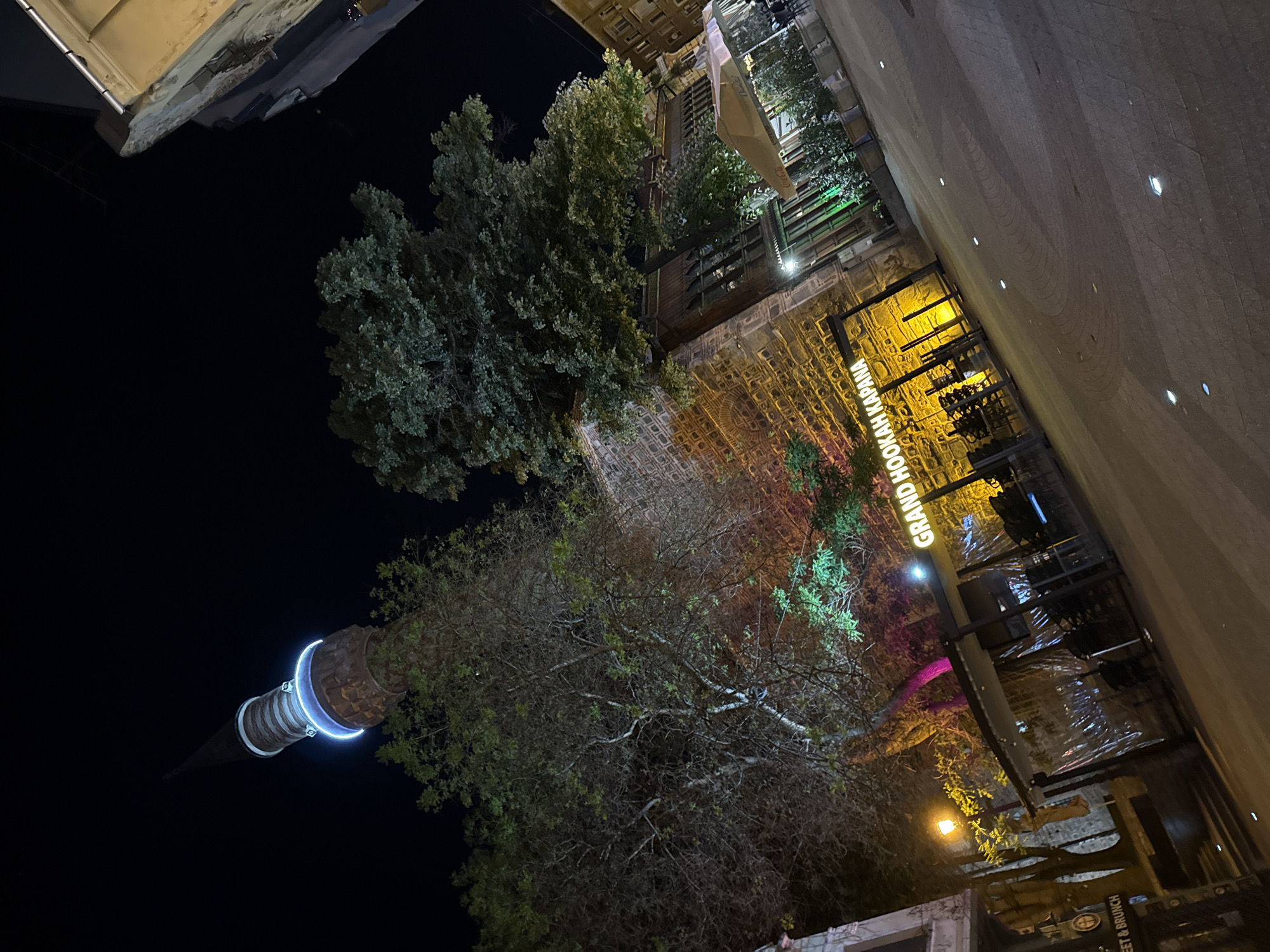 Captured from a low angle at night, the image features a minaret of the Dzhumaya Mosque illuminated with blue lights against a dark sky. The mosque is located in the city of Plovdiv, Bulgaria. The image shows the tower rising to the left with trees below it. To the right are commercial establishments, including a "Grand Hookah Kapana" with yellow lighting, set against a brick wall. Sidewalk and street details are seen in the foreground, indicating an urban environment.