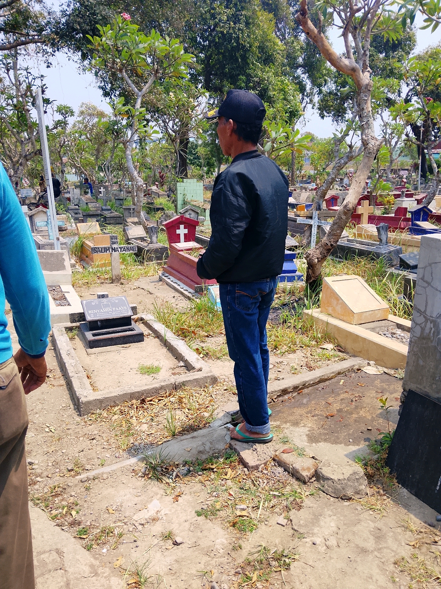 A man wearing a black cap, black jacket, and blue jeans stands in a cemetery in Kepuh, Indonesia. The cemetery is filled with gravestones and crosses of various sizes and materials, many of which are weathered and overgrown with grass and foliage. The ground is dirt and scattered with dry leaves. In the foreground, a person wearing a blue shirt and brown pants is partially visible. The man in the black jacket is looking away from the camera, towards the gravestones. The scene appears to be during daylight hours, with clear skies overhead.