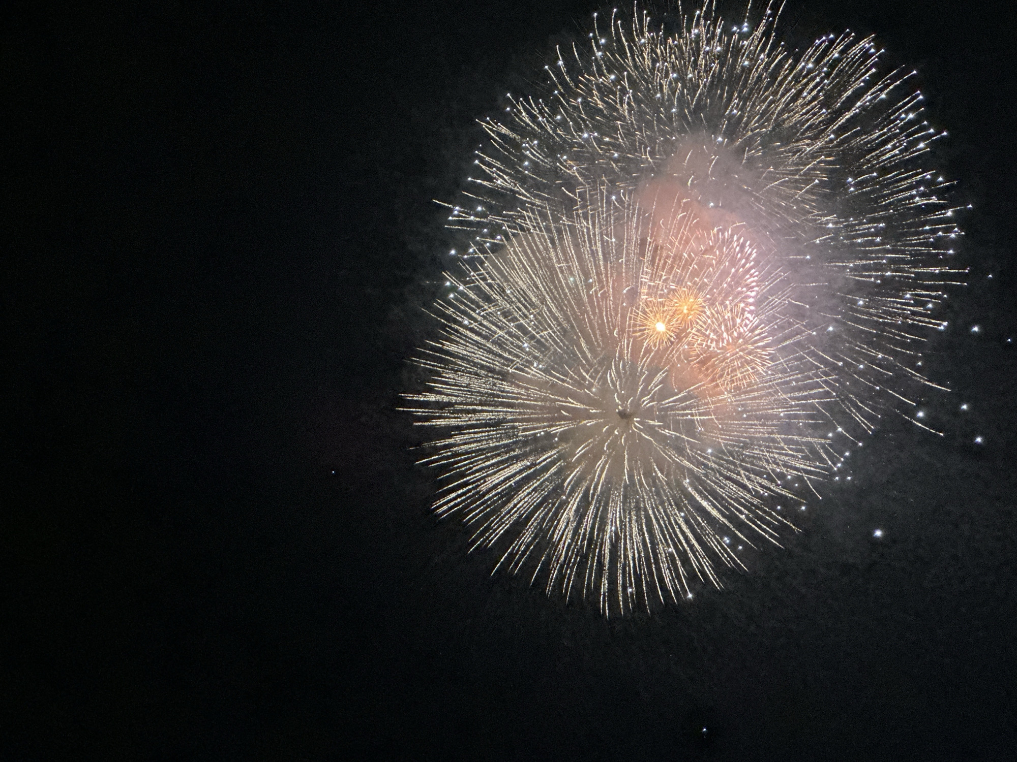 The image displays a vibrant fireworks display against a pitch-black night sky. The fireworks are primarily white and gold, with some hints of orange and pink at their core, resembling bursting dahlias. These pyrotechnic bursts are densely clustered in the lower right quadrant of the frame, with thin, luminous trails radiating outwards. The scene suggests an outdoor event, likely a celebration or festival. No specific landmarks are visible to pinpoint a location within Đà Nẵng, Vietnam, though the dark sky and fireworks are consistent with a nighttime event in any urban or coastal area.