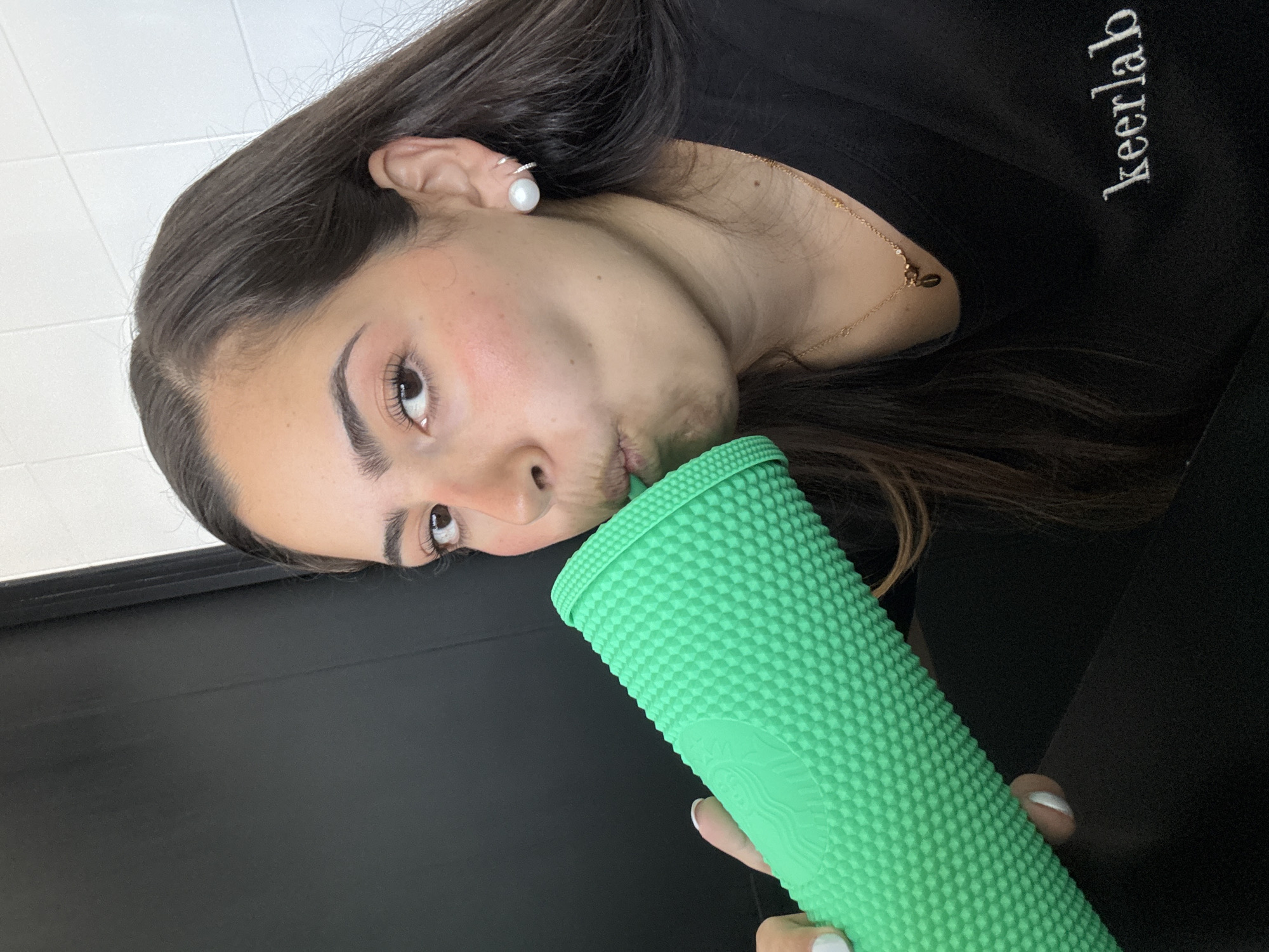 The image, taken with a tilted perspective, captures a young woman from the chest up, engaged in drinking from a bright green, studded Starbucks tumbler. Her head is tilted to the side, and her cheeks are slightly puffed as she sips. She has long, dark brown hair that flows around her shoulders, and her eyes, which appear brown, look towards the viewer. She wears a black top or sweatshirt, on which the word "keerlab" is embroidered in white capital letters on the right side of her chest.
Notable accessories include a white pearl stud earring with a small silver hoop just above it in her left ear, and a thin gold necklace with a small charm around her neck. Her fingernails are painted with white nail polish, visible on the hand holding the tumbler. The tumbler itself is a distinctive bright green with a textured, studded surface, and the Starbucks siren logo is subtly embossed on its side.
The background suggests an indoor setting, with a white tiled wall visible behind her head and a dark, possibly black, flat surface (like a table or counter) in the lower part of the frame. The overall lighting appears to be ambient indoor light, suggesting daytime. The woman's expression is neutral, focused on drinking. This scene takes place in San Bernardino Tlaxcalancingo, Mexico.