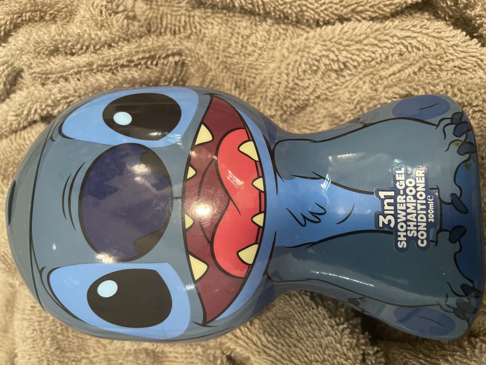 The image shows a close-up of a blue plastic bottle shaped like the character Stitch from Disney's Lilo & Stitch. The bottle is standing upright on a textured, beige towel. The Stitch character is depicted with large black eyes with blue irises, a wide open mouth showing red gums and sharp, yellow teeth, and blue fur details. Text on the bottle reads "3in1 SHOWER-GEL SHAMPOO CONDITIONER 300mle". The lighting suggests an indoor setting, possibly a bathroom. There is no indication of the city of Sevlievo, Bulgaria, or any people or specific actions occurring in the scene.