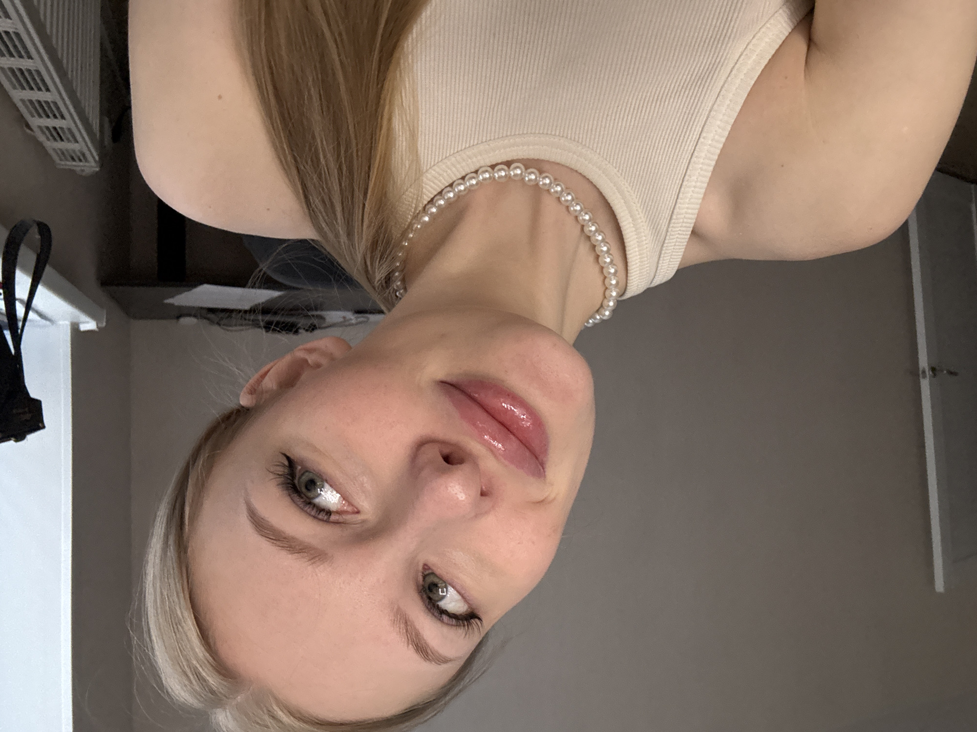 The image is a medium shot of a young, fair-skinned woman with blonde hair, taken indoors. She is looking directly at the camera with a playful expression, tilting her head slightly to the right. Her makeup includes noticeable mascara and lip gloss. She is wearing a cream-colored tank top and a pearl necklace. The background is a neutral-colored wall, with a dark desk and a radiator visible to the right. A white door is partially visible on the left. The image quality is slightly grainy. There is no information within the image to suggest it is located in Perm, Russia.