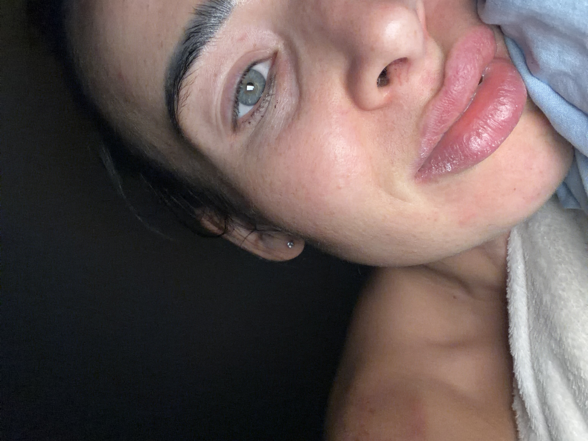A close-up portrait of a woman with light skin, gray-blue eyes, and dark eyebrows. She has a small earring in her left ear and appears to be leaning her head slightly to the side. A blue cloth is visible near her mouth, and a white towel is draped over her shoulder. The background is dark. The image does not contain enough information to determine the location in the city of Saint Petersburg, Russia.