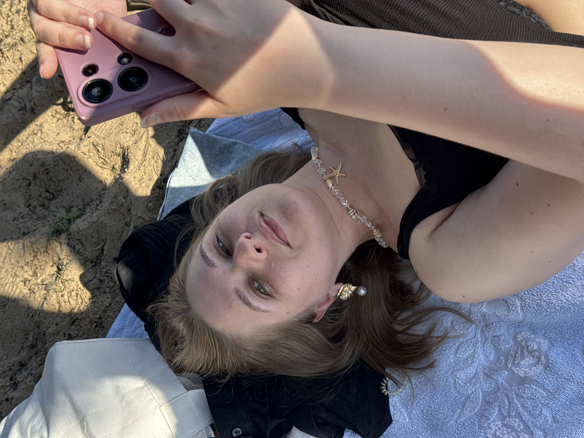 In this close-up, overhead shot, a young person with light brown hair is lying on their back on a towel, outdoors. They are holding a purple smartphone up, seemingly taking a selfie. Their face is angled upwards, looking towards the camera. They are wearing a black top and a necklace with clear beads and a starfish charm. They also have a pearl and gold earring in their visible ear. The background appears to be sandy, with some shadows cast by what looks like dried grass or twigs. The lighting suggests it might be daytime, possibly late afternoon given the warm tones and shadows. The overall mood seems relaxed and casual, possibly a beach or park setting.