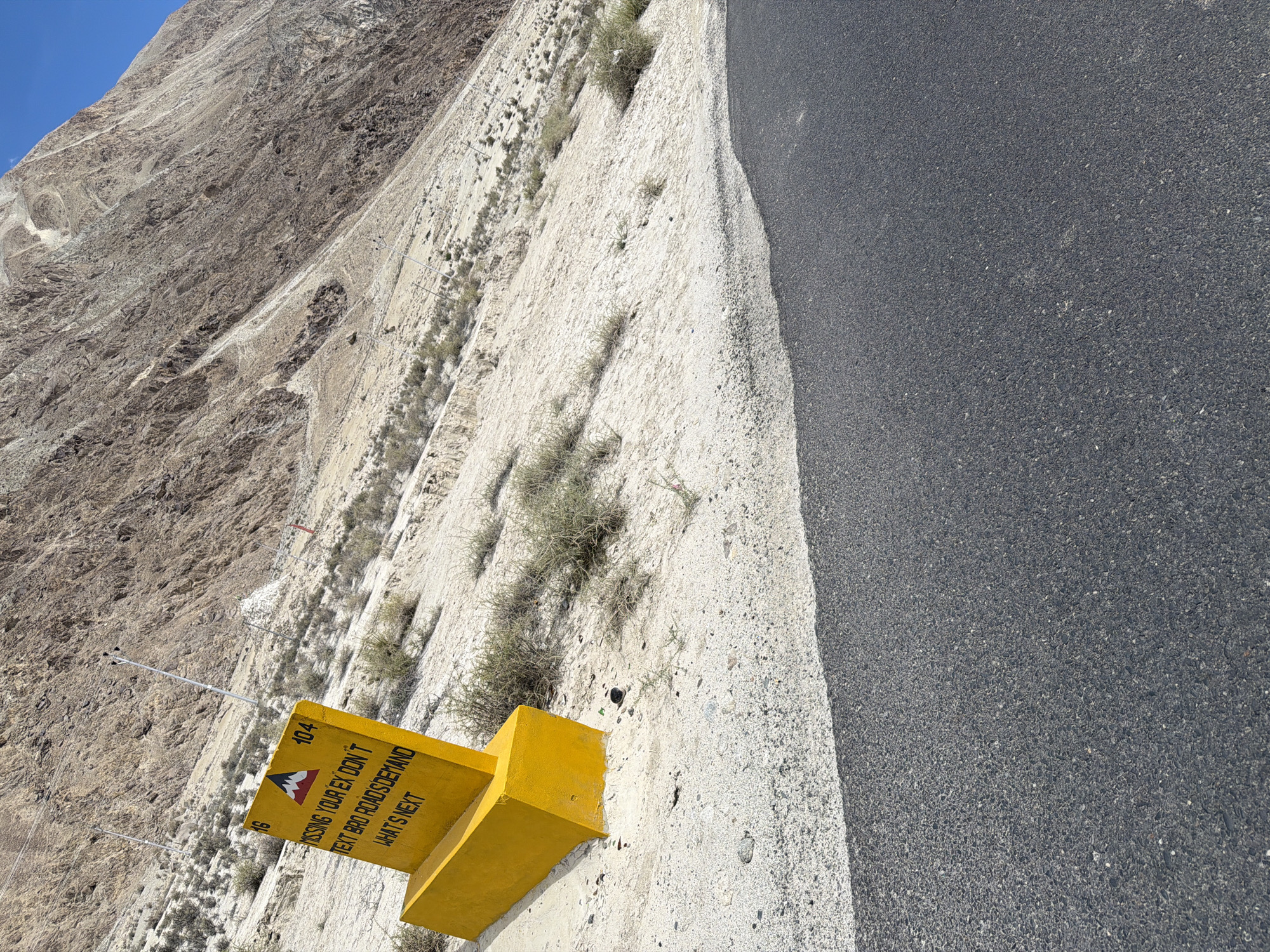 A yellow concrete post with black text is situated in a barren, mountainous landscape in Panamik, India. The post is positioned next to a paved road and has text that reads "MISSING YOUR EX DON'T TEXT BRO ROADS DEMAND WHAT'S NEXT". The mountains are rocky and dry, with sparse vegetation and a clear blue sky above. The road is dark asphalt with a white stripe along the edge.