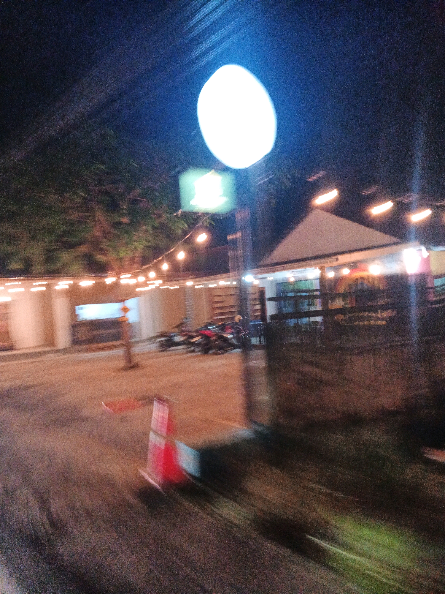 The image depicts a nighttime scene, likely in a commercial area of Kroya, Indonesia, given the context. A large, bright, out-of-focus circular light source dominates the upper portion of the image, casting a strong glow. Below this, a string of small, warm-toned lights hangs, illuminating a covered outdoor seating area or a pathway in front of a building. The building itself appears to be a single-story structure with a sloped roof.
In the middle ground, a red and white traffic cone is visible on the ground, suggesting a temporary obstruction or guidance marker. Further back, several motorcycles are parked or are in motion, indicated by their blurry forms. The ground appears to be paved or a hard surface, possibly a road or parking area. The overall impression is of a bustling but slightly chaotic scene, with motion blur suggesting movement, possibly from a vehicle the photo was taken from. Trees and foliage are visible in the background, adding to the outdoor ambiance. The atmosphere is dark, typical of nighttime, but punctuated by the artificial lighting.