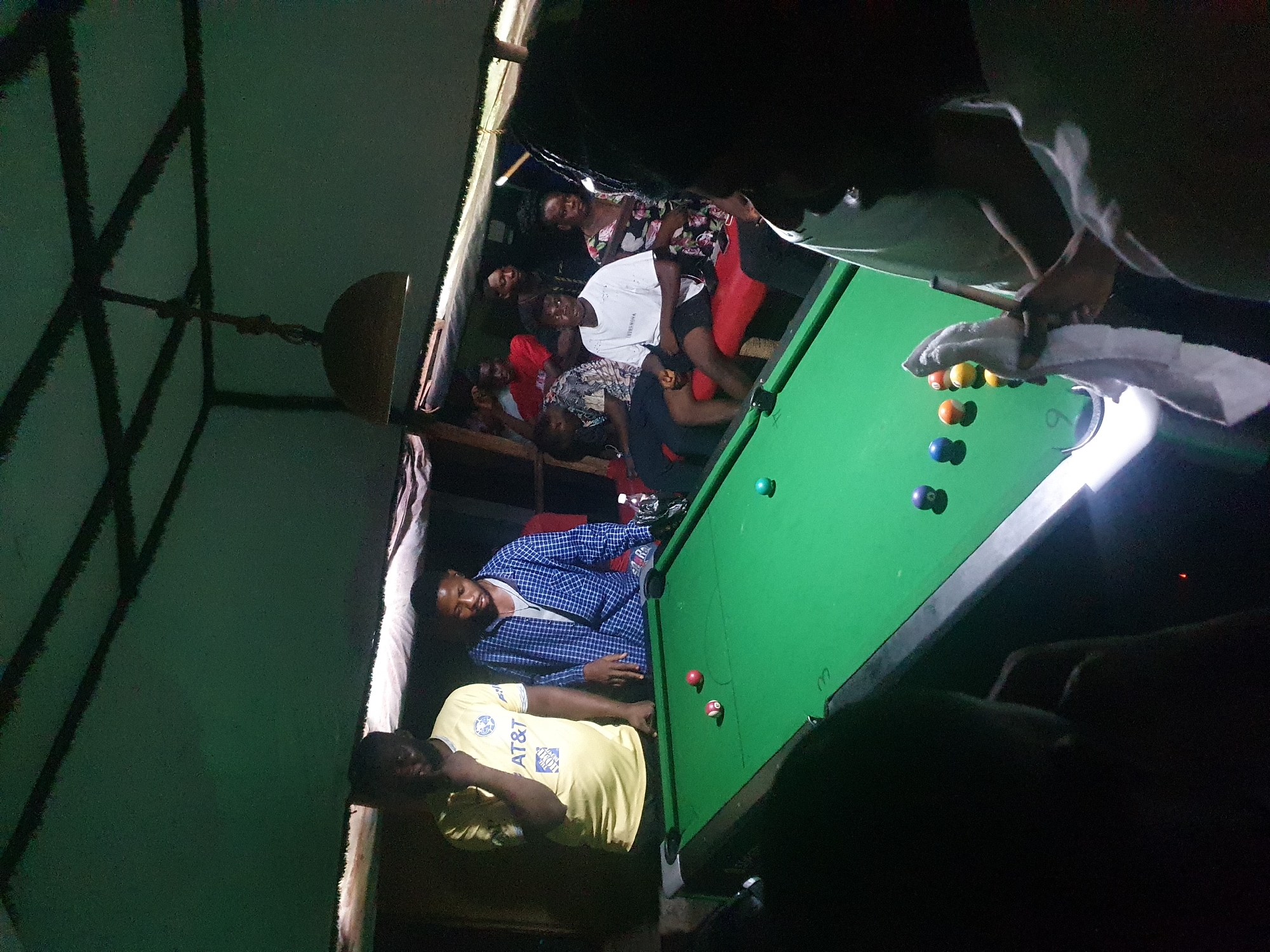 This image captures a vibrant social gathering in Lokoja, Nigeria, centered around a game of pool. The scene is set indoors at what appears to be a casual bar or game room, characterized by a rustic interior with dark, crisscrossing ceiling beams. The overall lighting is dim, contrasting with a bright overhead light that illuminates the green felt of the pool table.
Multiple individuals are present, engaged in playing or observing the game. One person is actively holding a pool cue, positioned to take a shot, while others are standing or sitting around the table, watching the action. In the foreground, a man in a yellow t-shirt, clearly displaying the "AT&T" and "HOME DEPOT" logos, stands beside another man wearing a blue checkered shirt. In the background, several other people are visible, some smiling towards the camera, including one person in a white t-shirt with "YAMAGATA" visible text.
The green pool table is equipped with an assortment of colored balls, including orange, blue, red, yellow, and black ones, with numbers "3" and "8" visible on some. A white towel is draped over the edge of the pool table. The atmosphere appears relaxed and convivial, indicative of friends enjoying a leisure activity during the nighttime.