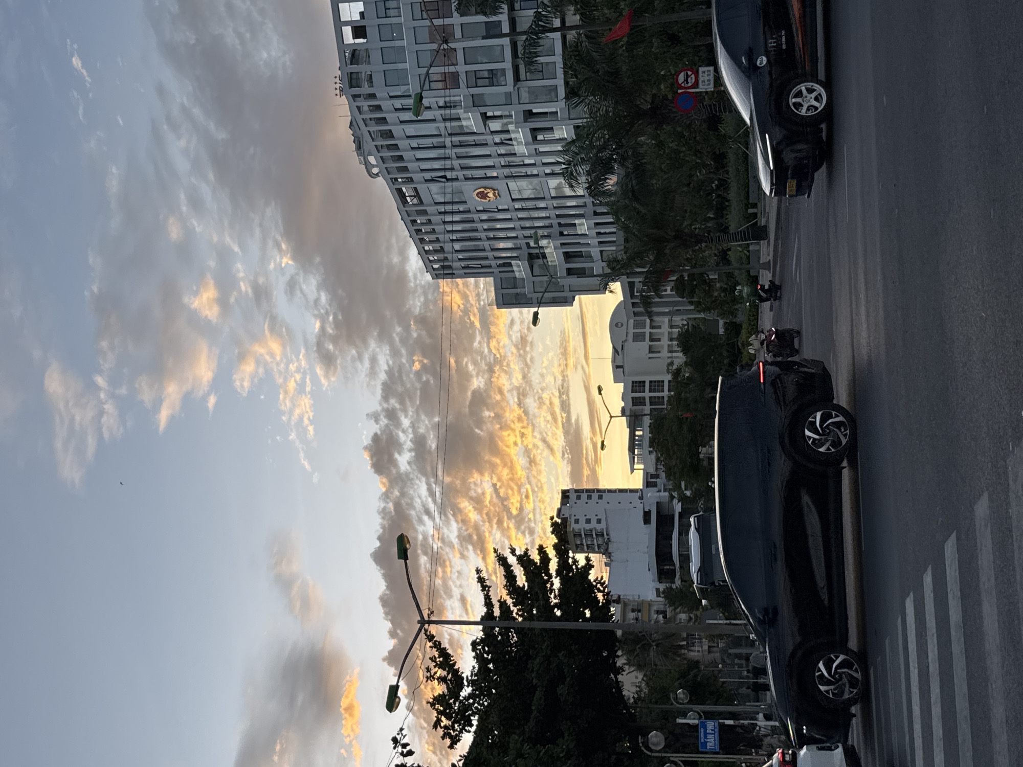 The image captures an urban scene in Nha Trang, Vietnam, at dusk. The sky is filled with dramatic clouds tinged with orange and yellow from the setting sun. In the foreground, a black SUV is stopped at a pedestrian crossing on a wide asphalt road. The vehicle has distinctive multi-spoke wheels. To the right, another black car, possibly a taxi, is in motion, showing a partial logo on its side. In the background, a modern, multi-story building with a patterned facade dominates the right side of the frame. A circular emblem, possibly a crest or logo, is visible on the building's exterior. Palm trees and other lush green foliage are scattered throughout the scene, suggesting a tropical climate. Streetlights are illuminated, casting a soft glow. Several other vehicles and a few individuals on motorbikes are visible in the mid-ground, indicating active city life. A street sign reading "TRẦN PHÚ" is present on the left side of the image, identifying the location as Tran Phu street, a prominent boulevard in Nha Trang.