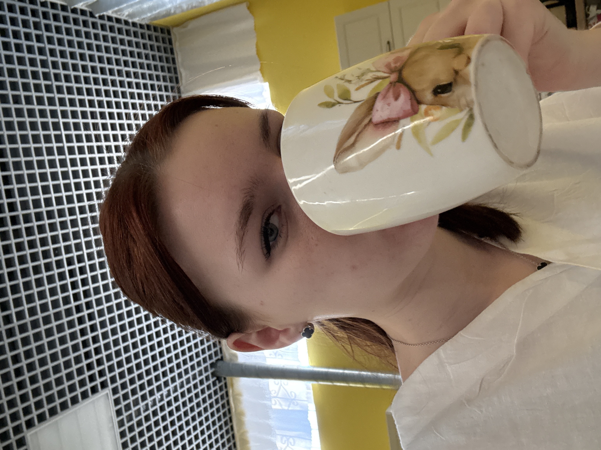 A woman drinks from a mug decorated with a bunny. She is wearing a white shirt and a delicate necklace. The background shows a wall of small, black square tiles and a kitchen with yellow walls. The exact location in Saint Petersburg, Russia cannot be determined from the image.