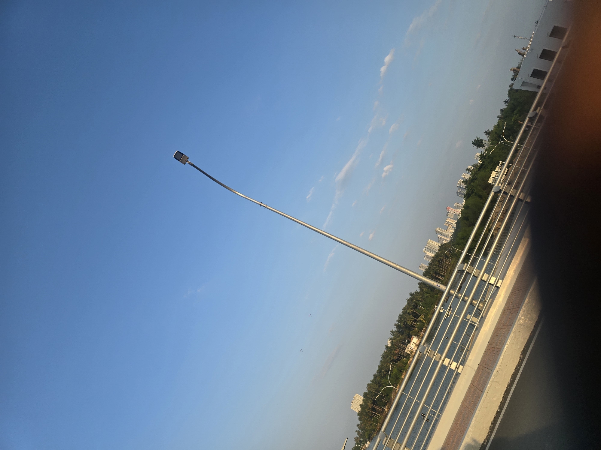 The image shows a view from a bridge in Nha Trang, Vietnam. The sky is a clear blue with a few wispy clouds. A tall, silver street lamp curves over the bridge. In the distance, there is a body of water, lined with lush green palm trees and some buildings. Across the water, a city skyline is visible with several tall buildings. The bridge has a silver railing and a paved walkway. The lower portion of the image is dark, suggesting it might be a reflection or a shadow.