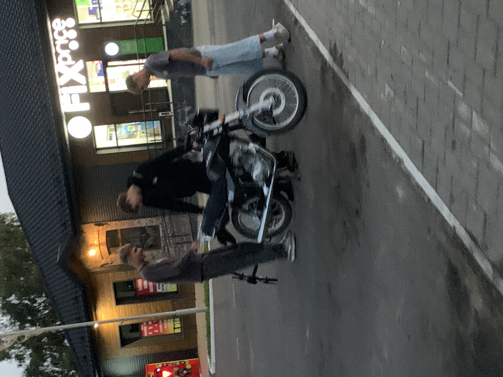 The image depicts three young men standing around a motorcycle in front of a "Fix Price" store. The store appears to be located in the city of Kapyĺ, Belarus, as indicated by the store's name and distinctive facade. One man, wearing a dark long-sleeved shirt and dark pants, is standing to the left of the motorcycle, looking down. Another man, in a black hoodie and dark pants, is positioned behind the motorcycle. The third man, wearing a grey t-shirt and light blue shorts, stands to the right of the motorcycle, looking down at it. The motorcycle itself is black with chrome accents and appears to be parked on an asphalt surface adjacent to a paved walkway. The background shows the "Fix Price" store with its illuminated signage and entrances, and a dark-tiled roof. The lighting suggests it is either early morning or late evening.