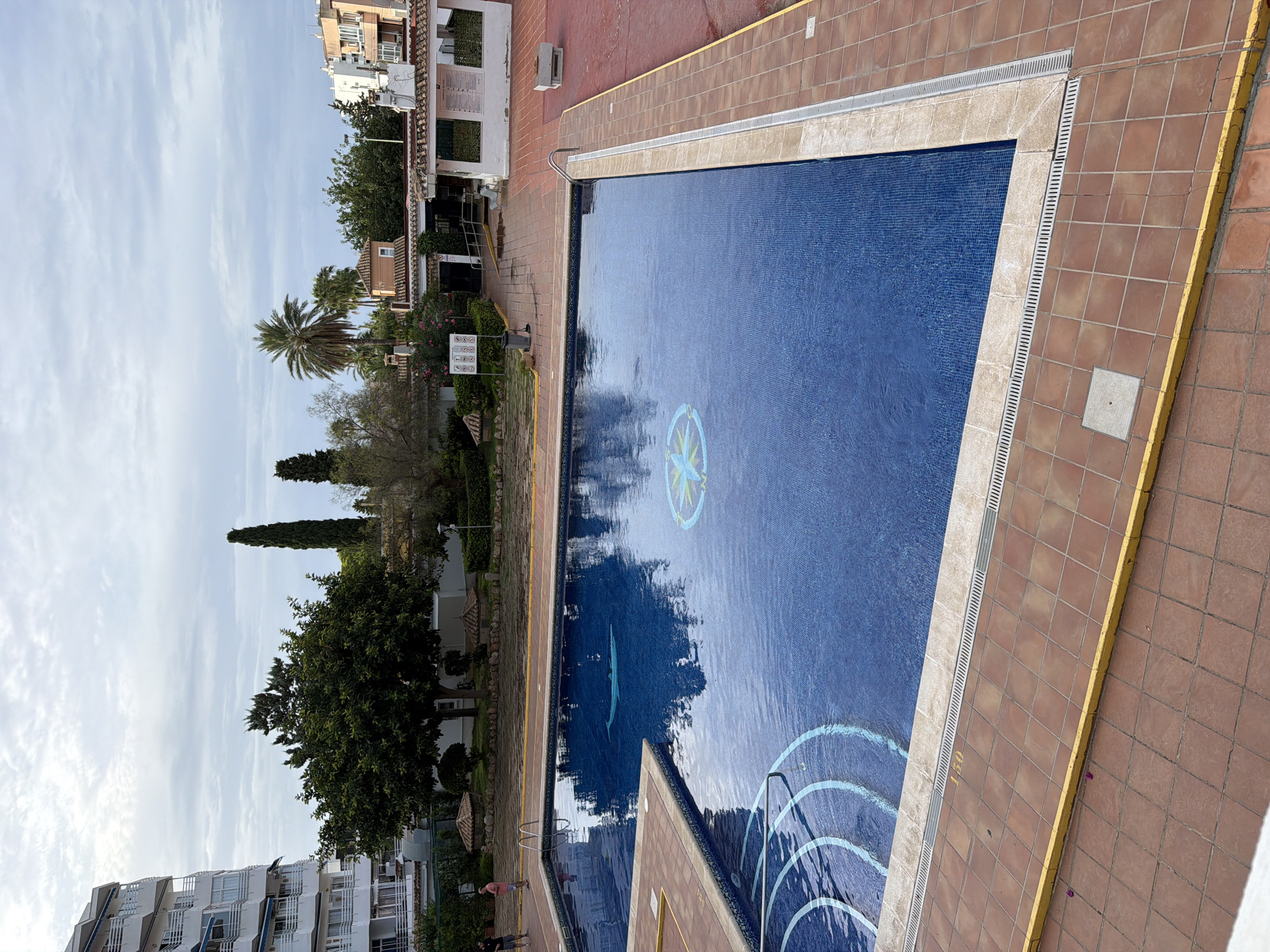 The image depicts a view of an outdoor swimming pool area in Palmanova, Spain, under a cloudy sky. The pool is rectangular with blue tiled mosaic surfacing and features a decorative compass rose design at the bottom. A section of steps with blue and white markings is visible on the left side of the pool. The pool is surrounded by a tiled patio area with terracotta-colored square tiles and a yellow border. In the background, there is a white building with multiple balconies and a building with a red-tiled roof, along with lush green trees and palm trees. Two men are standing on the patio to the left of the pool, facing away from the camera. The overall scene is a typical resort or residential complex setting.