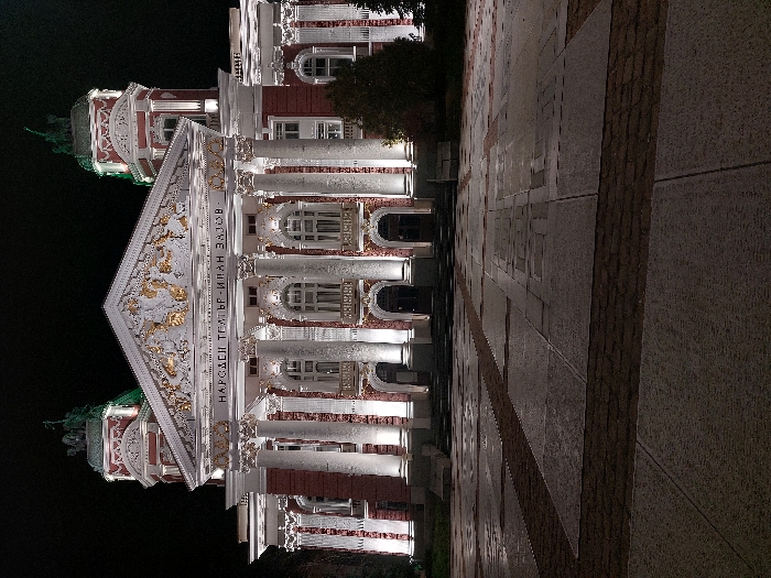 The image displays the illuminated facade of the Ivan Vazov National Theatre in Sofia, Bulgaria, at night. The grand neoclassical building features a red brick base with prominent white architectural details, including six fluted Corinthian columns supporting a triangular pediment. The pediment is adorned with golden and white sculptural reliefs, and below it, the golden inscription "НАРОДЕН ТЕАТЪР ИВАН ВАЗОВ" is visible. Two ornate corner towers, each topped with a cupola and a sculptural group, flank the main structure. White spotlights illuminate the theatre's columns, details, and the statues on the towers, with a subtle green light emanating from the tower cupolas. The foreground consists of a vast, unpopulated plaza paved with a mosaic of grey flagstones and darker reddish-brown cobblestone strips, appearing wet and reflective, suggesting recent rain. The sky above is dark and clear, characteristic of nighttime. No individuals, vehicles, or significant activity are present in the scene.