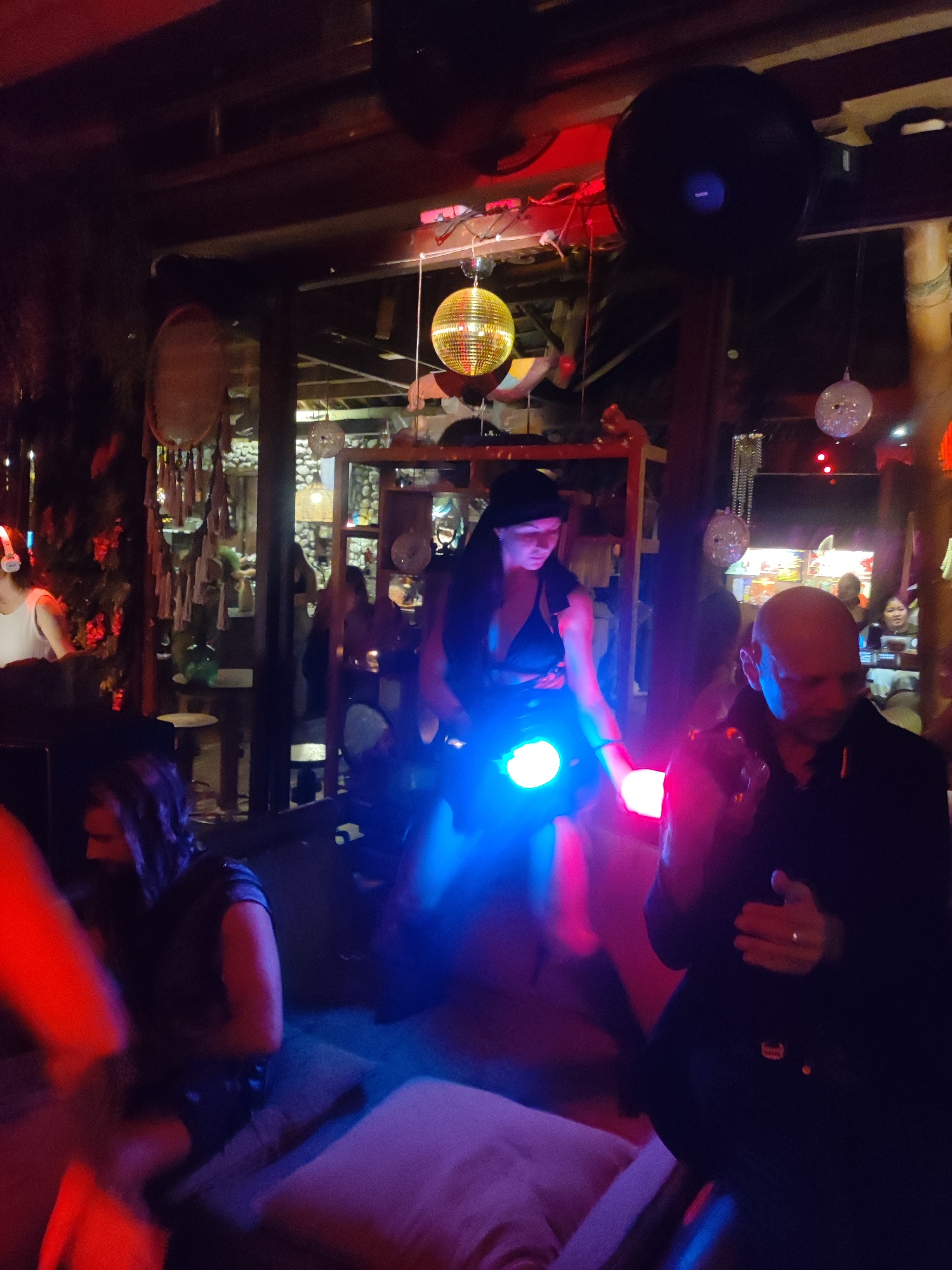 The image depicts a dimly lit interior, possibly a bar or club, in Lokaserena, Indonesia. The lighting is a mix of ambient and artificial, with a disco ball and colored lights casting blue and red hues. Several people are visible, including a woman with a black beanie holding illuminated objects (likely light sticks), a bald man in a dark jacket gesturing, and a person with long hair wearing a sleeveless top. In the background, other patrons are seated or standing, and a decorated shelf or display case is visible. The environment suggests a relaxed, social gathering in an enclosed space.