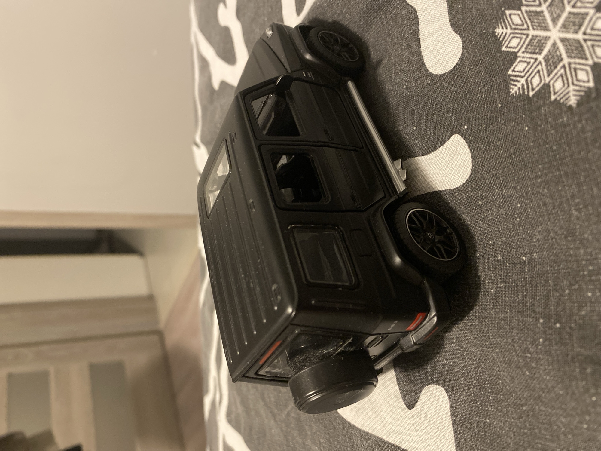 A black toy Mercedes-Benz G-Class is positioned on a gray and white patterned surface. The toy car is viewed from a three-quarter rear angle, showing its spare tire mounted on the back, tinted windows, and a sunroof. The surface it rests on appears to be a textile, possibly a blanket or tablecloth, with a repeating pattern of stylized snowflakes or abstract shapes. In the background, out of focus, are elements of an indoor environment, including what appear to be doorways or furniture with neutral-colored surfaces. There is no discernible indication of the city of Łódź, Poland, or any specific location.