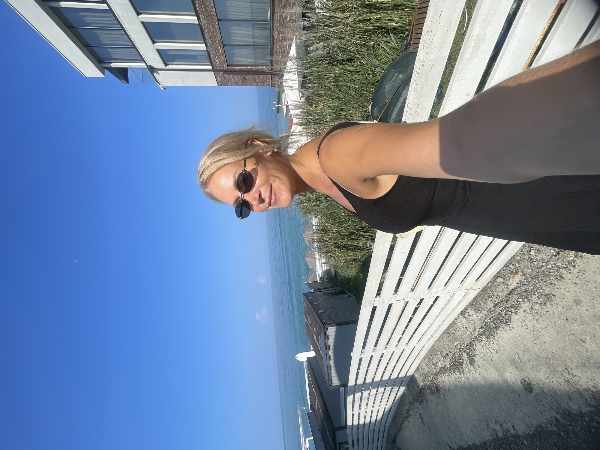 A woman with blonde hair, wearing sunglasses and a black dress, is taking a selfie outdoors. She is standing next to a white wooden fence, with a view of the sea and buildings in the background. The sky is clear blue with a faint crescent moon visible. The overall setting appears to be a coastal area in Sochi, Russia, possibly a resort or residential complex, given the presence of modern architecture and beach umbrellas. The sunlight suggests it is daytime.