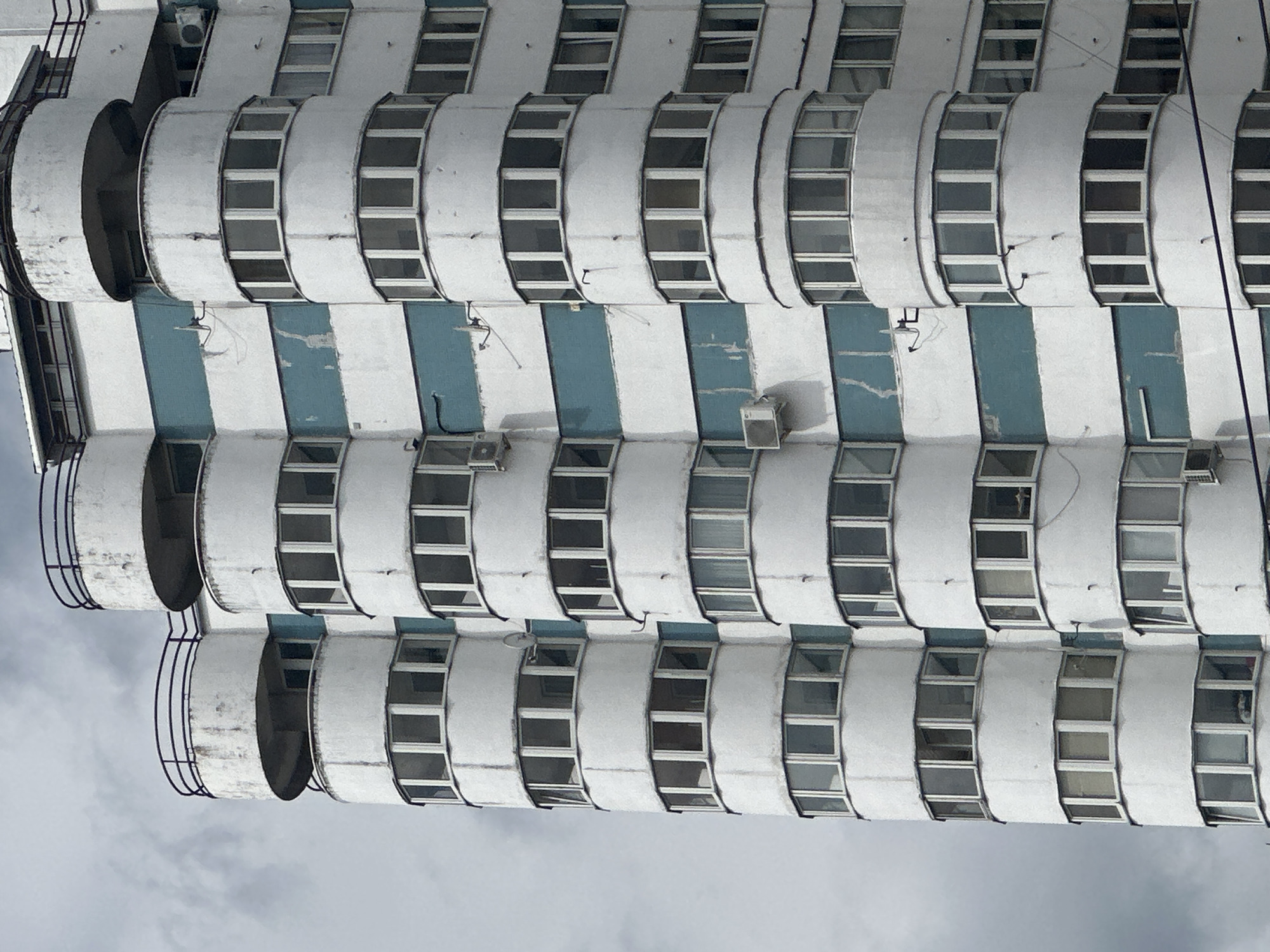 An image, presented as if rotated to its upright position, captures the facade of a multi-story building from a low-angle perspective, looking upwards towards an overcast sky. The architecture features a repetitive, modular design dominated by rows of curved, semicircular balcony or bay window structures, stacked vertically. Each curved section is fitted with multiple narrow, rectangular windows that form a continuous band along its face.
The building's exterior is primarily white, with noticeable vertical stripes of light blue or teal color running down its length, separating the stacks of curved balconies. The white surfaces, especially on the balconies, show signs of weathering, including dirt streaks, discolored patches, and some minor deterioration, suggesting the building's age. Several small air conditioning units are affixed to the blue-painted sections of the wall. Some dark metal railings are visible on the outermost balconies towards the left of the frame.
The windows generally appear dark, reflecting the light from the sky or the dim interiors. No people are visible, and no specific activities are taking place. The overall impression is one of stark, functional architecture, characteristic of some Soviet-era or post-Soviet designs commonly found in cities like Minsk, Belarus, which aligns with the provided location context. The sky is a uniform, light grey, indicating an overcast day. No discernible text is visible on the building.