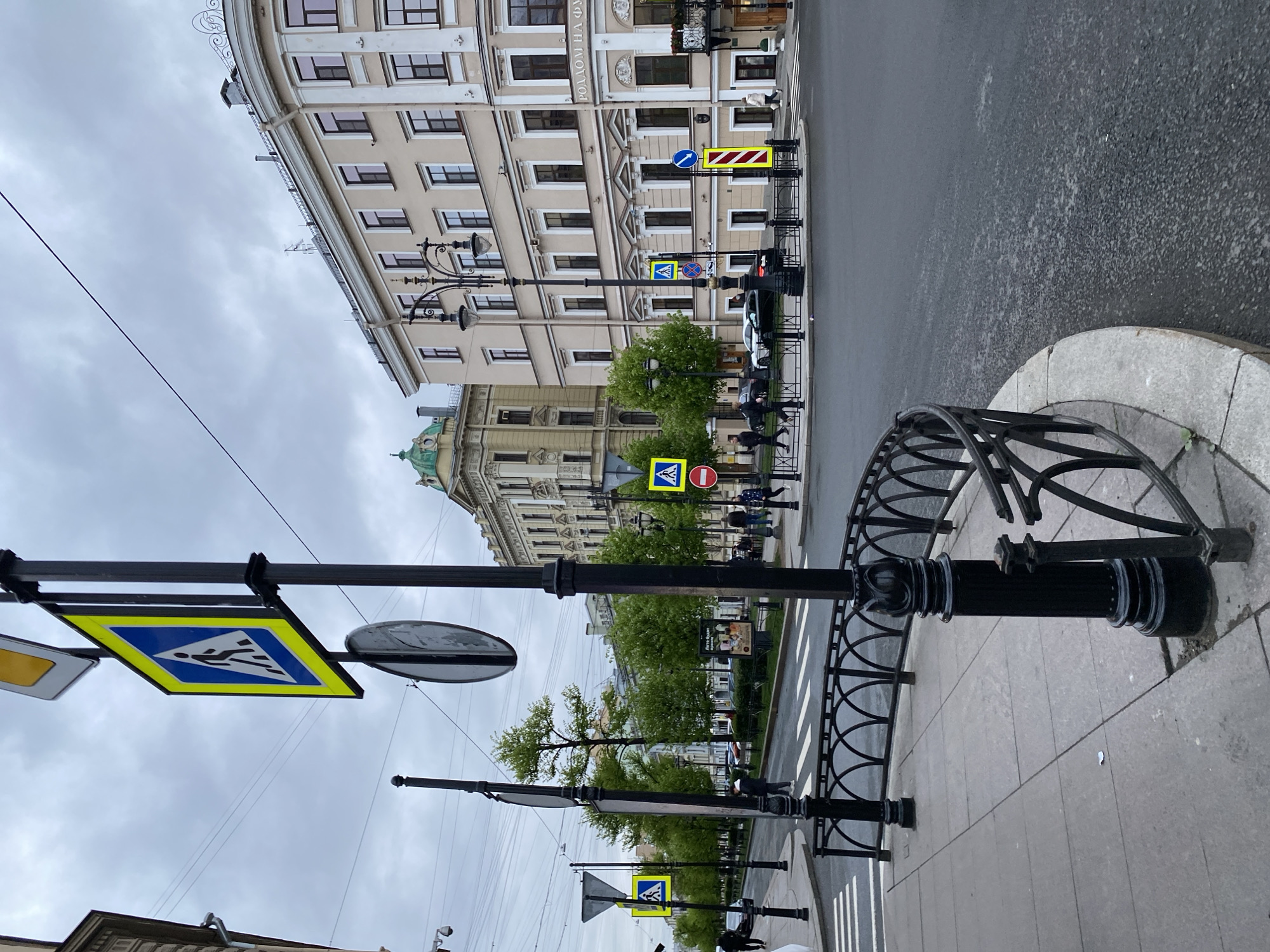 The image depicts a street scene in Saint Petersburg, Russia, characterized by overcast skies and damp pavement. The foreground features a pedestrian walkway with black metal fencing and decorative posts. Several yellow and blue pedestrian crossing signs are prominently displayed, affixed to black poles. The signs are slightly angled. In the midground, the street is visible with crosswalk markings. Cars and pedestrians can be seen moving along the street. Buildings, classic of Saint Petersburg architecture, line the street. One of the buildings bears the inscription "РОДОМ НА ФУ..." in Russian. Lampposts are also visible.