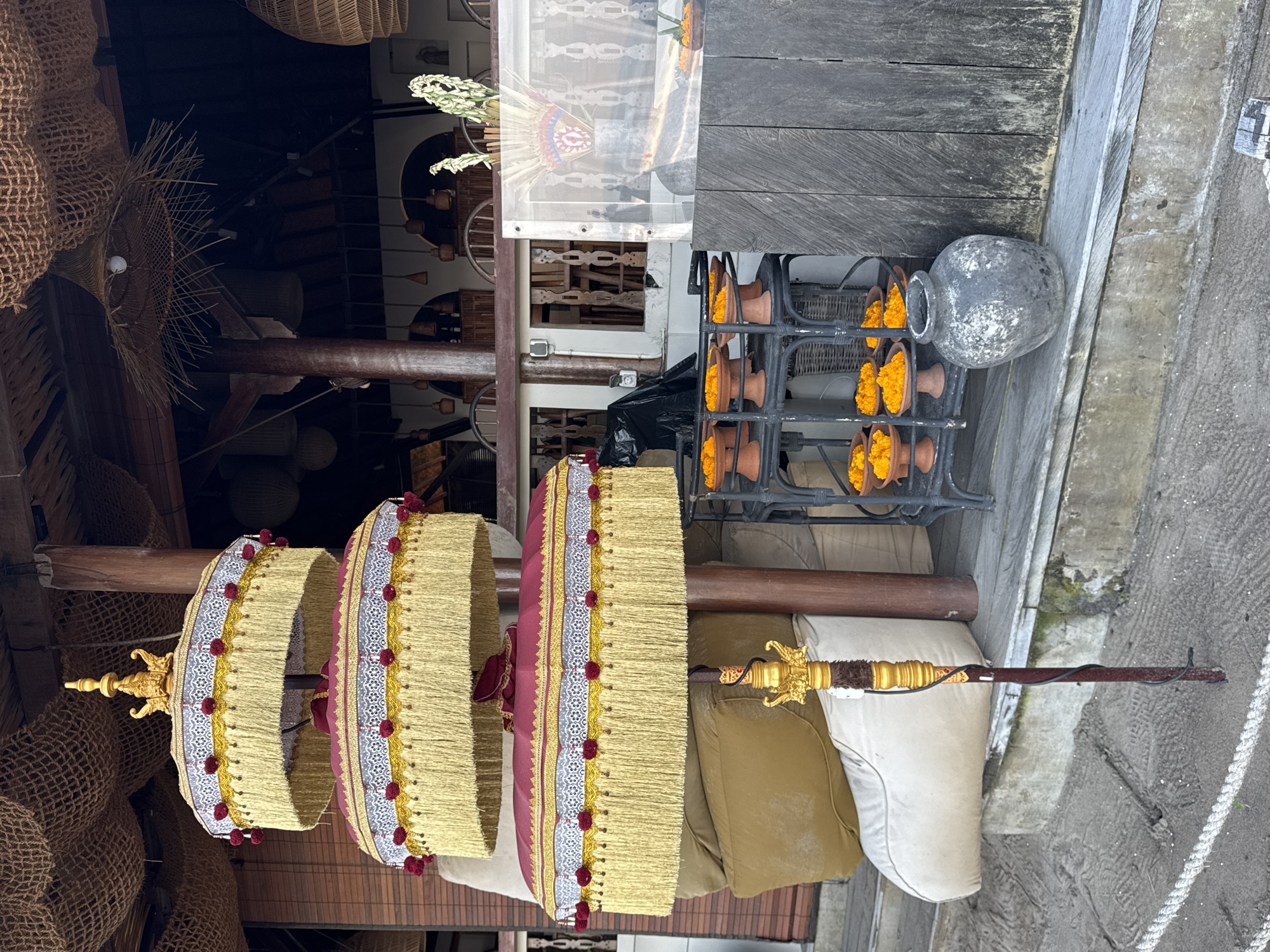 The image, taken in Kuta, Indonesia, depicts a vibrant, multi-tiered ceremonial umbrella (likely a Balinese *Tedung*) standing prominently in the foreground. The umbrella features three large, circular tiers, each adorned with a gold-fringed edge, intricate white and gold patterned fabric, and a line of red pom-poms or tassels. The central pole is dark brown, topped with an ornate gold finial and an elaborate golden section further down.
To the right of the umbrella, a metal rack holds multiple small, terracotta pots, each brimming with bright orange flowers, likely marigolds used for offerings. Next to this rack, a weathered, grey ceramic or stone pot rests on the ground.
The background on the left reveals the interior of what appears to be a shop or market stall, filled with various woven goods, including hanging basket-like objects and textured netting. Wooden beams and structures are also visible. On the right, dark wooden planks form a wall or counter, and a light-colored, possibly concrete or stone, ground is visible with a braided rope lying on it. Cushions, one beige and one off-white, are placed near the base of the umbrella pole.
The overall scene suggests a cultural display or preparation, possibly for a religious ceremony or a decorative element for a business, with the *Tedung* and flower offerings being strong indicators of Balinese traditions. The setting appears to be outdoors or in an open-air structure during daylight hours. No people or visible text are present.