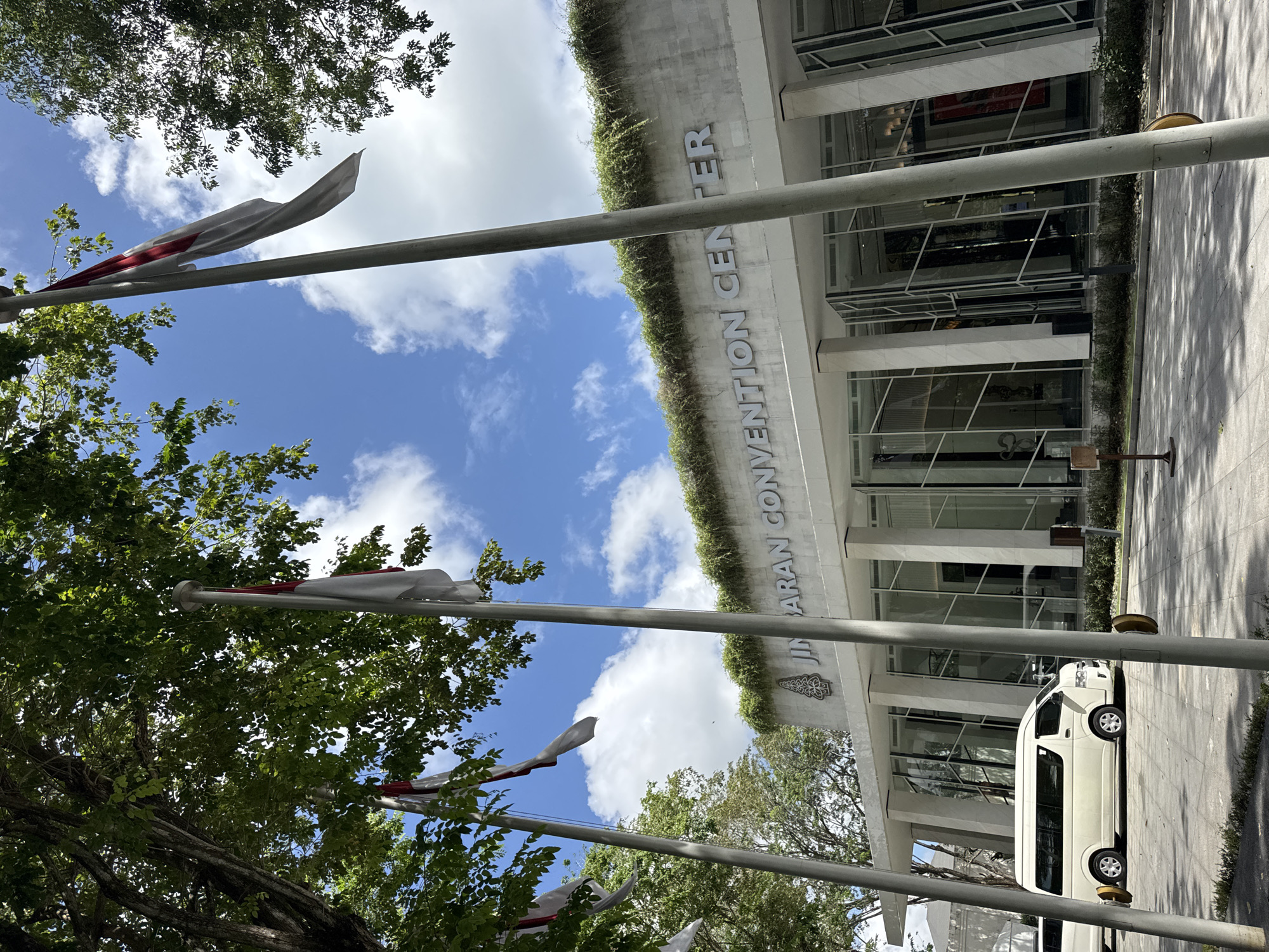 The image shows the front entrance of the JICA (Japan International Cooperation Agency) Convention Center in Kuta Selatan, Indonesia. The building is a modern, white structure with large windows and the convention center name displayed prominently on the facade. Foliage is growing on the roof of the building. Several flagpoles with Indonesian flags are visible in the foreground, along with trees and blue sky with clouds. A white van is parked on the right side of the frame. The scene is shot from a low angle, tilting upwards.