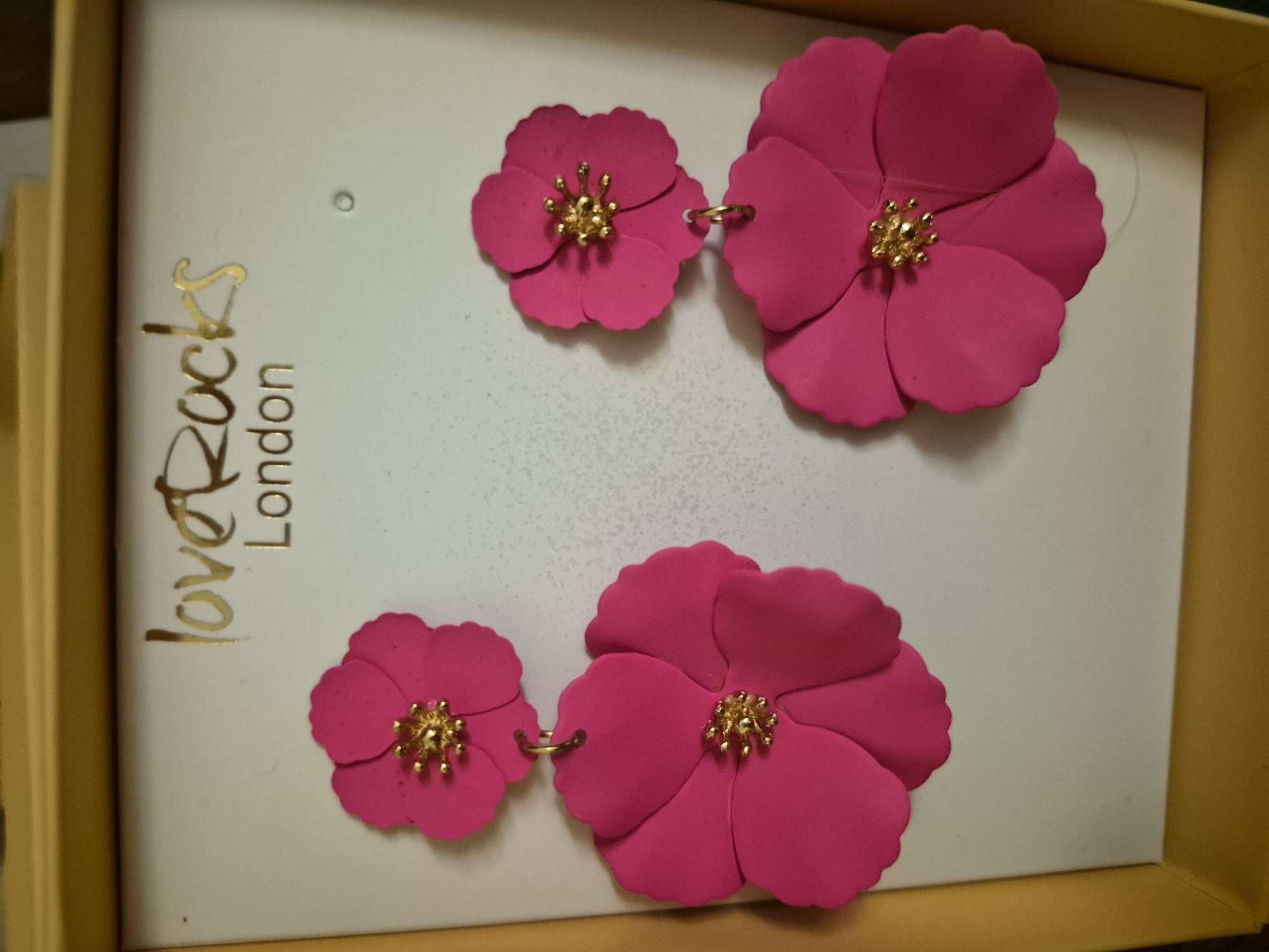 In a yellow box, a pair of bright pink flower-shaped earrings with gold centers are displayed against a white background. The earrings are a statement piece, with each earring featuring two flower shapes stacked on top of each other. The larger flower is at the bottom, with a smaller flower above it. The "loveRocks London" logo is visible in gold lettering at the top of the white display. The overall scene is bright and cheerful, suggesting the earrings are a fun and fashionable accessory.