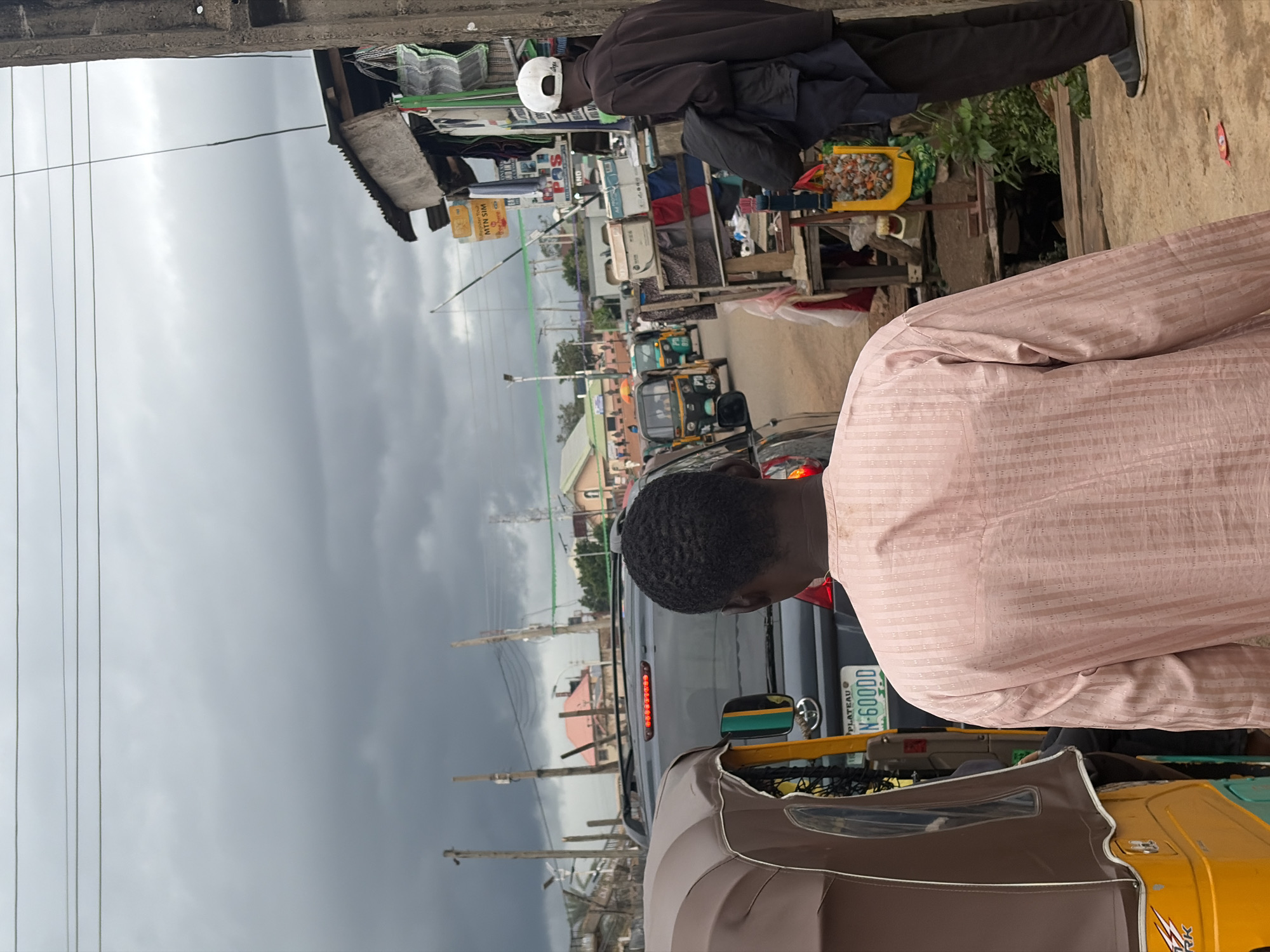 The image depicts a street scene in Gura Top, Nigeria, under a cloudy sky. In the foreground, a man wearing a light pink patterned shirt is seen from behind, walking away from the camera. To his left, the side of a brown and yellow auto-rickshaw is visible. Further down the street, a dark colored SUV with its brake lights illuminated is stopped in traffic. Next to the SUV, another auto-rickshaw with a green body is visible. On the right side of the street, there is a makeshift stall or shop with various items displayed, including what appear to be clothes, bags, and boxes. A man wearing a dark jacket and a white cap stands near the stall, looking to his right. The street is lined with poles and wires, suggesting an urban or semi-urban environment. The overall impression is one of a busy street with ongoing traffic and commercial activity.