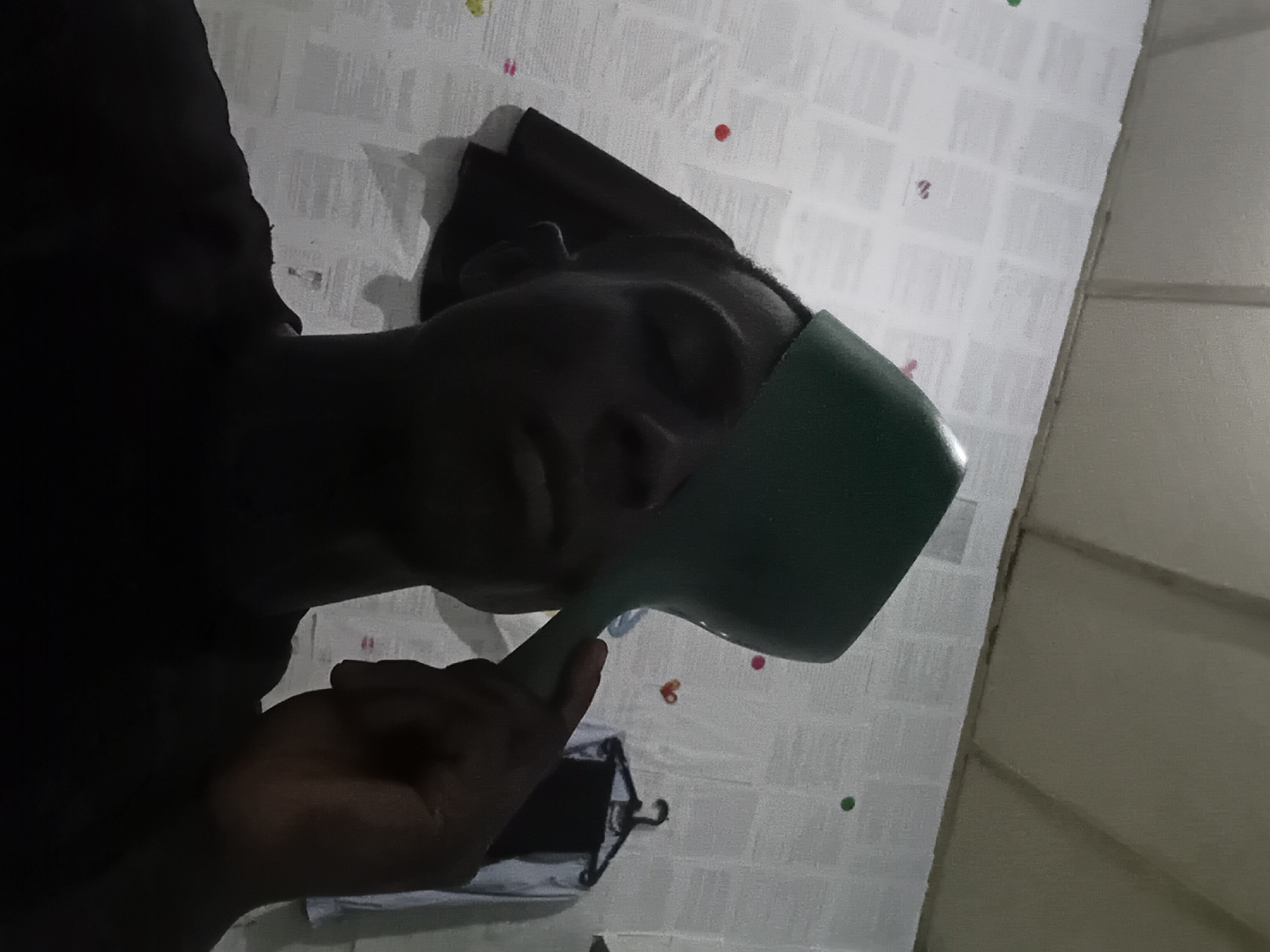 A man with dark skin is holding a green plastic cup to his face. He is wearing a dark-colored shirt or robe. The background is a white wall with a grid pattern and small colored dots. A clothes hanger with a dark garment is visible near the man's hand. The scene appears to be indoors. There is no explicit evidence within the image to confirm its location in Akwete, Nigeria.