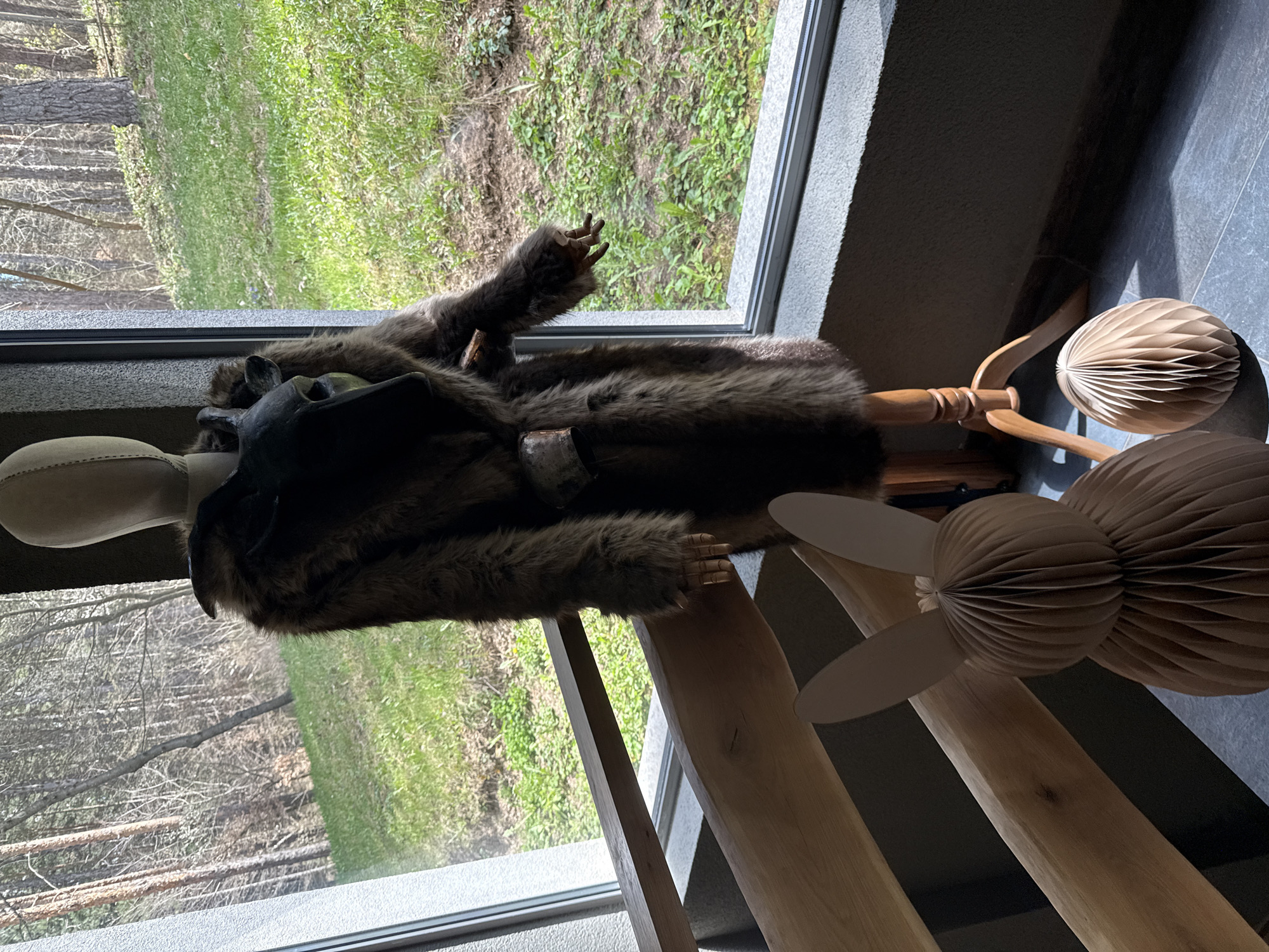 The image shows a room with a window overlooking a grassy area with trees. A mannequin stands in front of the window wearing a fur coat and a black vest. There is a decorative paper fan on a wooden stand near the mannequin. The location is Velingrad, Bulgaria.