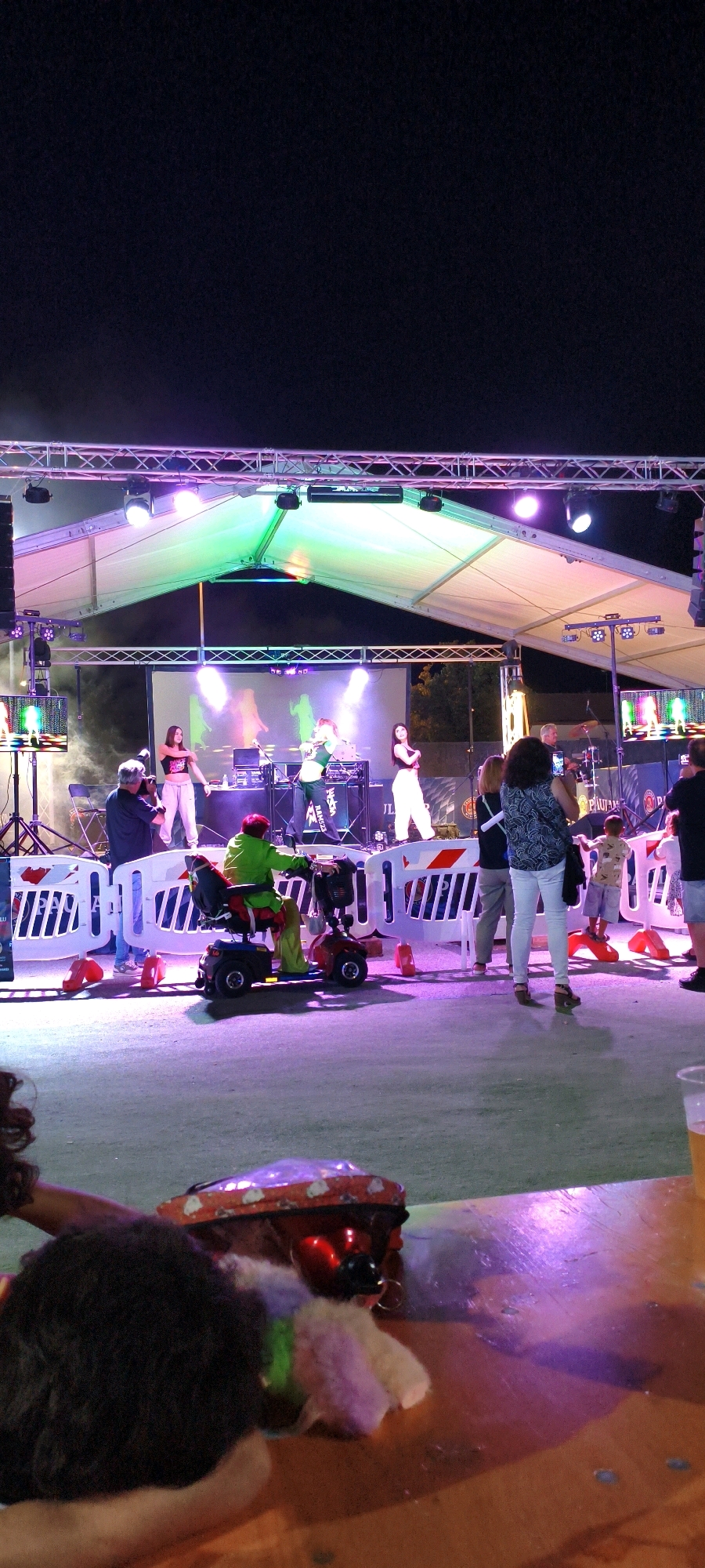 An outdoor evening event is depicted, with a stage set up for a performance. On stage, there are several people, appearing to be performers, with musical equipment and a large screen displaying silhouettes of dancers. The stage is illuminated by colorful lighting, primarily green and white. In front of the stage, a crowd is gathered, watching the performance.
In the foreground, to the left, a person is partially visible, with dark, curly hair and wearing a black top. They are seated at a wooden table that appears to be part of an outdoor seating area. On the table, there is a red object with reflective elements and what appears to be some colorful, fluffy material. To the right of this person, there is a blurry view of a glass containing a yellowish liquid, likely a beverage.
To the right of the stage, a person in a green outfit is seated on a red and blue mobility scooter, facing the stage. Other individuals are visible in the crowd, some standing and observing, while others are interacting. The ground appears to be a paved surface, illuminated by stage lights and overhead lighting.
The overall atmosphere suggests a lively event with music and entertainment taking place at night. The location is confirmed as Campiglia Marittima, Italy.
