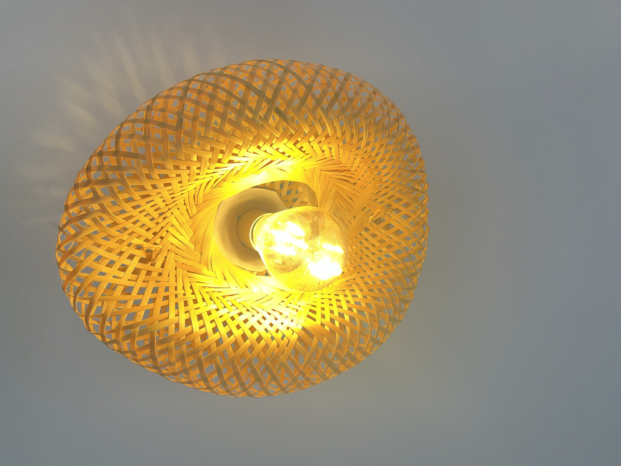 This is a close-up, eye-level shot of a woven bamboo lampshade with a lit lightbulb inside, hanging against a plain gray background. The lampshade is circular in shape and intricately woven, creating a patterned effect with open spaces. Light emanates from the bulb, casting a warm yellow glow and creating a halo effect around the light source. The woven structure of the lampshade allows light to filter through, projecting subtle, starburst-like patterns onto the wall in the upper left quadrant of the image. The lightbulb itself appears to be an incandescent bulb, visible within the center of the lampshade. The overall impression is one of simple, natural design and warm, inviting illumination. The background is a neutral gray, which helps the yellow light and the natural tones of the bamboo to stand out. There is no text visible in the image. The context of Abuja, Nigeria, does not provide specific visual cues within this image itself, as it is a close-up of an interior object.