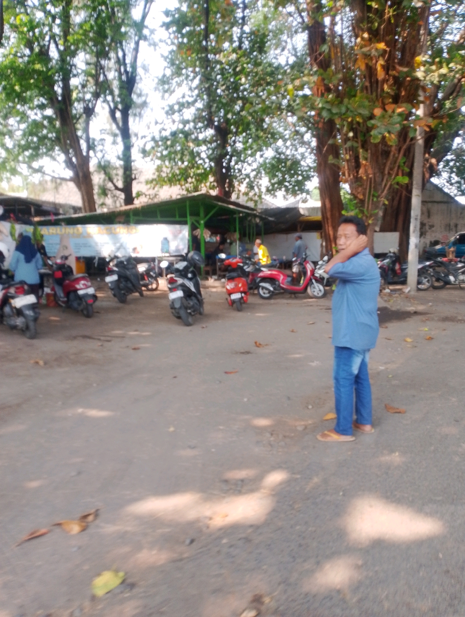 The image depicts an outdoor scene in what appears to be Probolinggo, Indonesia, likely a parking area or marketplace with a small food stall. In the foreground, a man wearing a blue shirt and blue jeans stands with his right hand touching the back of his neck, looking towards the right of the frame. He is wearing light-colored sandals. Behind him, several motorbikes are parked. To the left, a woman in a blue headscarf stands near a red motorbike. A green-roofed structure, possibly a food stall with signage, is visible in the background, where other people and motorbikes can be seen. The ground is a dirt or gravel surface, with fallen leaves scattered around. Sunlight filters through the trees, casting shadows on the ground. The overall environment suggests a casual, open-air setting.