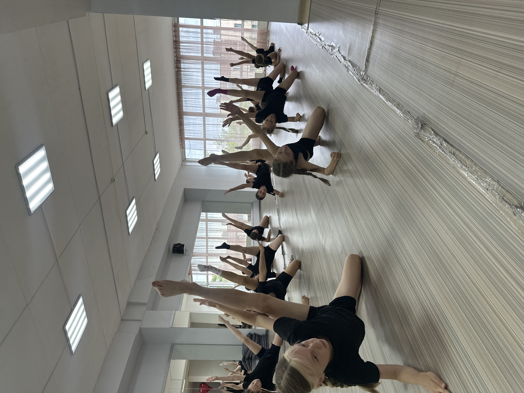 A group of young female dancers in black leotards and shorts are practicing ballet in a studio. They are positioned on a wooden floor in various stretching poses, with their legs extended in the air. The studio has large windows with light-colored curtains and mirrors on one wall, reflecting the dancers and the room. Fluorescent light fixtures are visible on the ceiling. The overall scene depicts a dance class in progress. The location in Votkinsk, Russia is not determinable from the image alone.