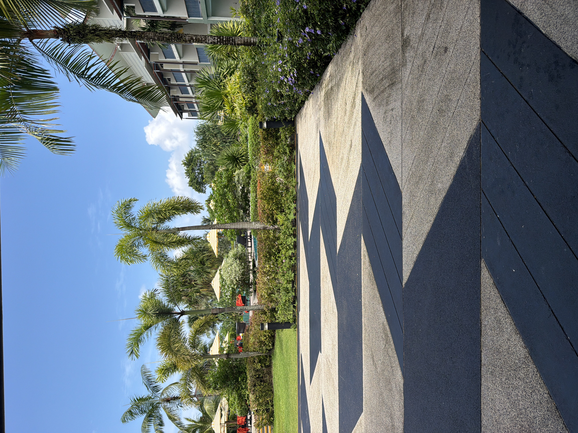 The image shows an outdoor area with a walkway in the foreground and a resort-like setting in the background. The walkway is paved with contrasting dark grey and light speckled concrete in a geometric pattern. Lush green vegetation, including palm trees and manicured bushes, borders the walkway. In the midground, there are lounge chairs and umbrellas, suggesting a pool area. A multi-story building with balconies is visible behind the greenery, characteristic of a hotel or resort. The sky is bright blue with scattered white clouds. The lighting indicates it is daytime, likely midday, with strong shadows cast by the walkway's pattern and the foliage. The environment appears to be tropical and well-maintained.