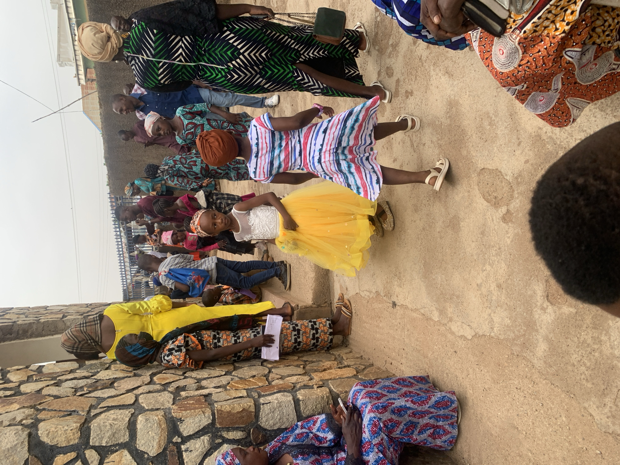 The image shows a gathering of people in an outdoor space, possibly in Jos, Nigeria. Several individuals are dressed in colorful African prints and fabrics. A young girl stands out in a white top and a bright yellow tulle skirt, while another girl wears a multi-colored striped dress. Some people are seated on what appears to be a stone wall, while others stand in a crowd. A building or structure is visible in the background, along with what appears to be a metal fence. The environment seems to be a meeting or event, with people engaging in conversation and observation. Some individuals are wearing head coverings or turbans.
