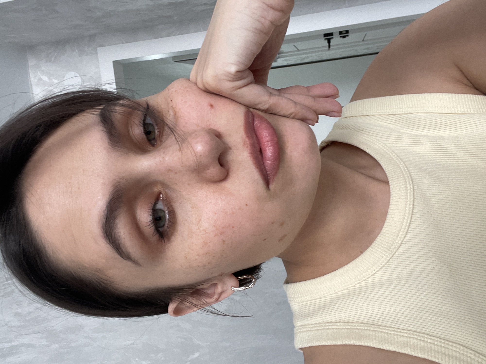 A close-up selfie shows a fair-skinned woman with dark hair and freckles. She is wearing a light yellow ribbed tank top and a small hoop earring in her left ear. Her left hand is partially covering the left side of her face, near her cheek. The background appears to be a bathroom with light gray walls and a mirror. No information indicates the location is in Izhevsk, Russia.