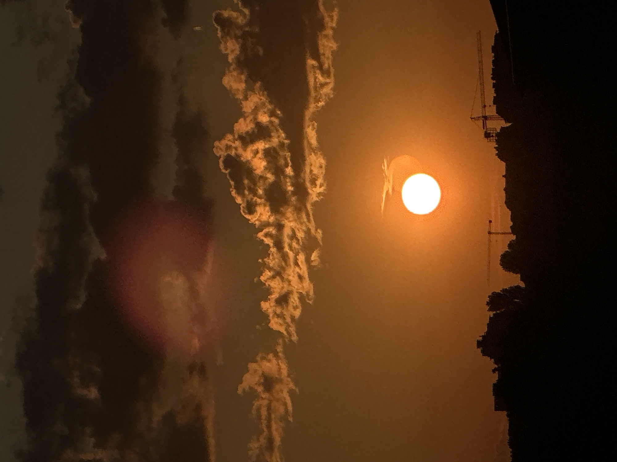 The image depicts an urban sunset over a developing area of Sofia, Bulgaria, characterized by a dominant orange and dark brown color palette. The sun, a brilliant white-yellow disk, is positioned low in the sky, slightly to the right of the vertical center, casting a warm glow across the entire frame. A prominent red circular lens flare artifact is visible in the upper left quadrant.
The sky is stratified into distinct layers. The uppermost section features dark, heavy cumulus clouds, primarily in shades of deep brown and gray, with their undersides subtly illuminated by the sunset's orange hue. Below these, a band of smaller, more defined clouds stretches horizontally across the mid-section, appearing bright orange and textured against the deeper orange sky. The lower sky, around the sun, is largely clear, displaying intense orange and amber tones that gradually darken towards the top. A minute white dot, possibly a distant star or planet, is faintly discernible in the far upper right corner.
The horizon is composed of a dark, silhouetted cityscape, indicative of an urban or suburban area within Sofia undergoing construction. Multiple multi-story buildings with varied rooflines form an irregular silhouette against the bright sunset. Interspersed among these structures are the dark, vertical forms of trees. Two significant construction cranes are visible against the horizon. A large, horizontal tower crane, its structure faintly outlined in yellow-orange, is prominent in the middle-right, extending over the silhouetted buildings. A second, smaller crane is partially visible on the far left, mostly obscured by a building silhouette. No people or animals are visible, and no discernible actions are taking place. The scene conveys a tranquil, yet industrially active, urban vista during dusk.