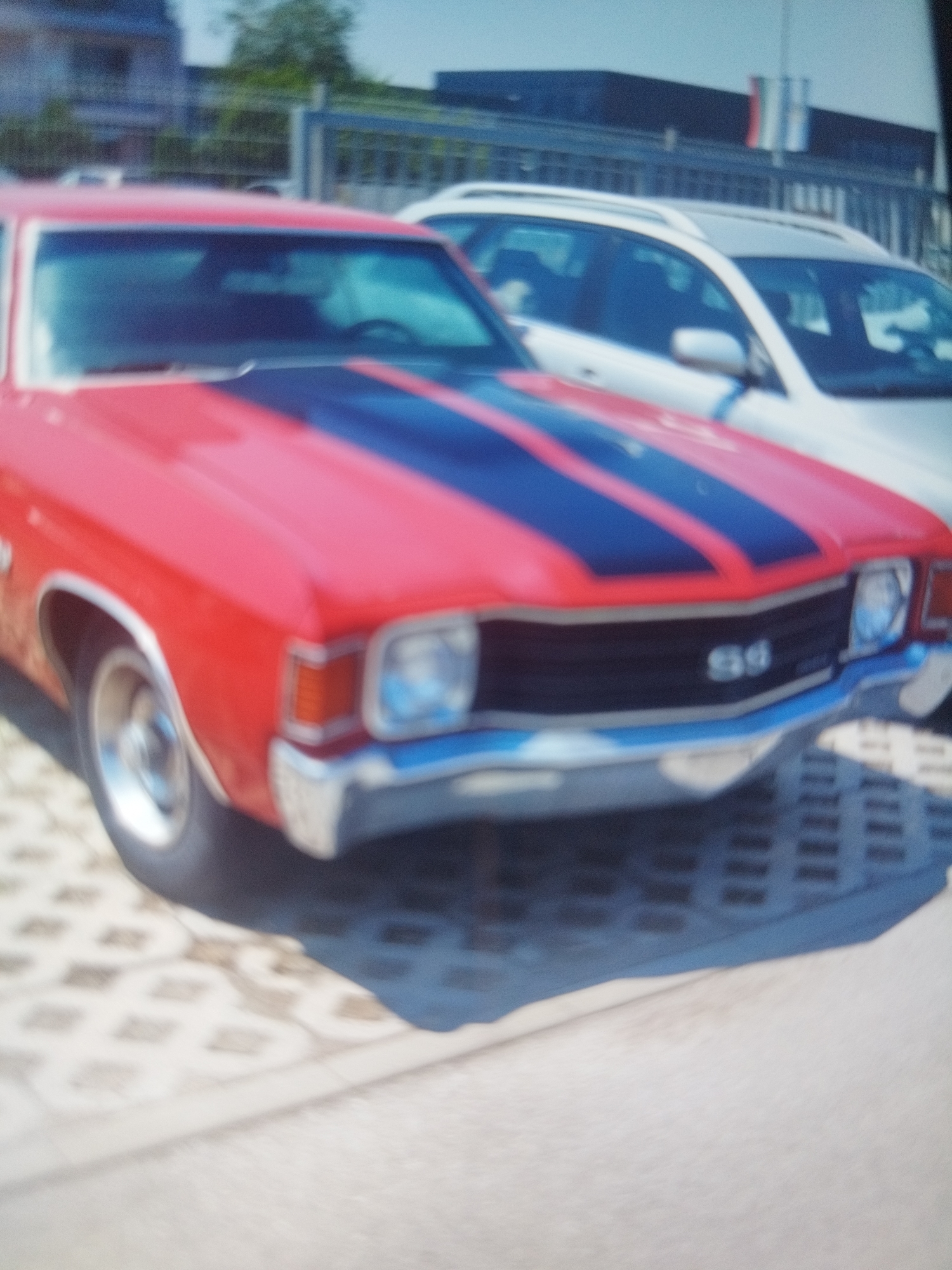The image depicts a red 1970-1972 Chevrolet Chevelle SS with black racing stripes parked on a concrete interlocking paver surface. The car is facing the viewer. A white or light-colored wagon or SUV is parked behind and slightly to the right of the Chevelle. In the background, there is a building and a fence or barrier with flags. Given the likely location in Plovdiv, Bulgaria, the building in the background might be associated with a commercial or industrial area. The overall image quality is blurry, making it difficult to discern finer details.
